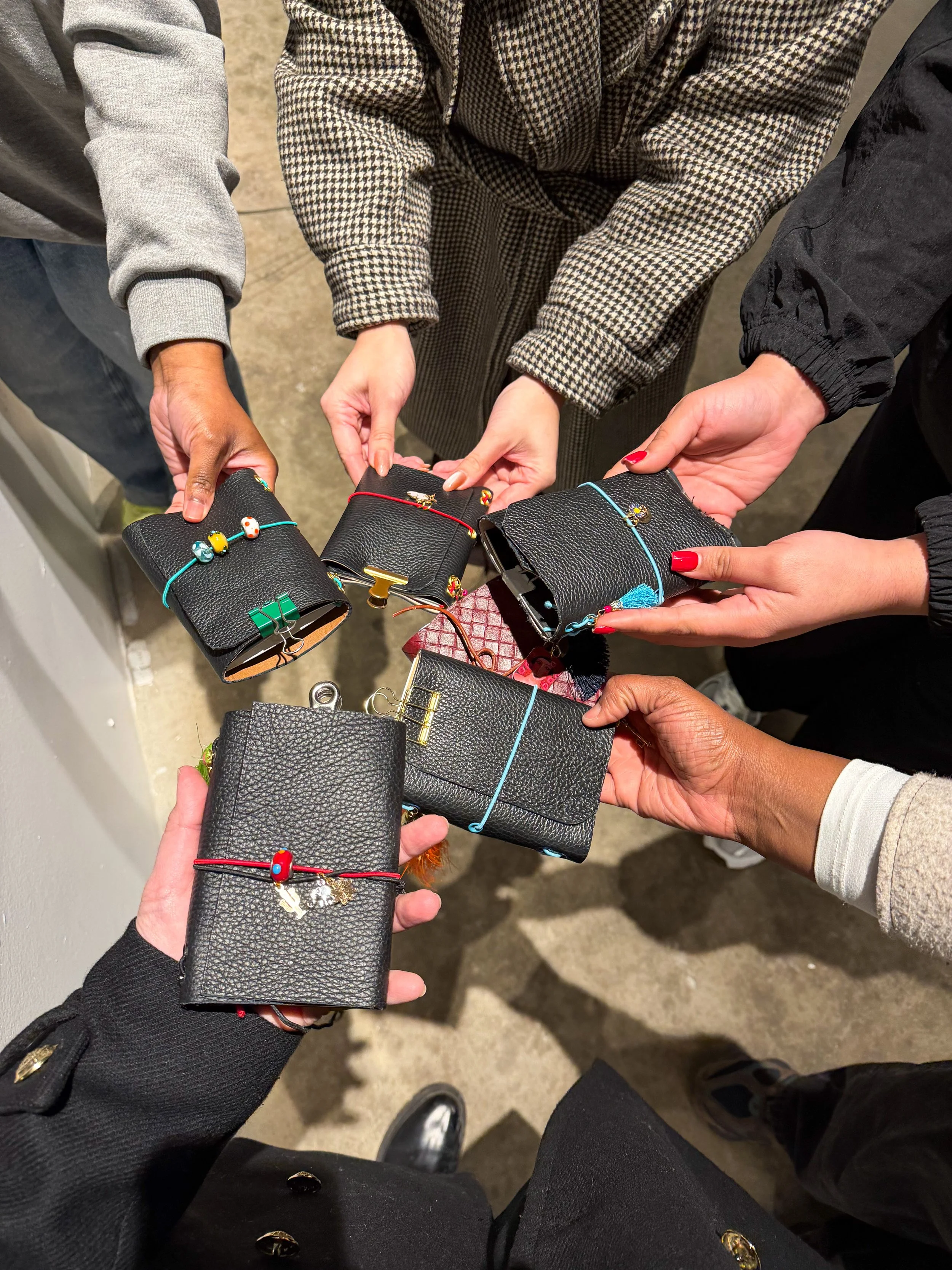 Several people holding black leather wallets or small handbags with colorful charms and accessories, displayed in a circle.