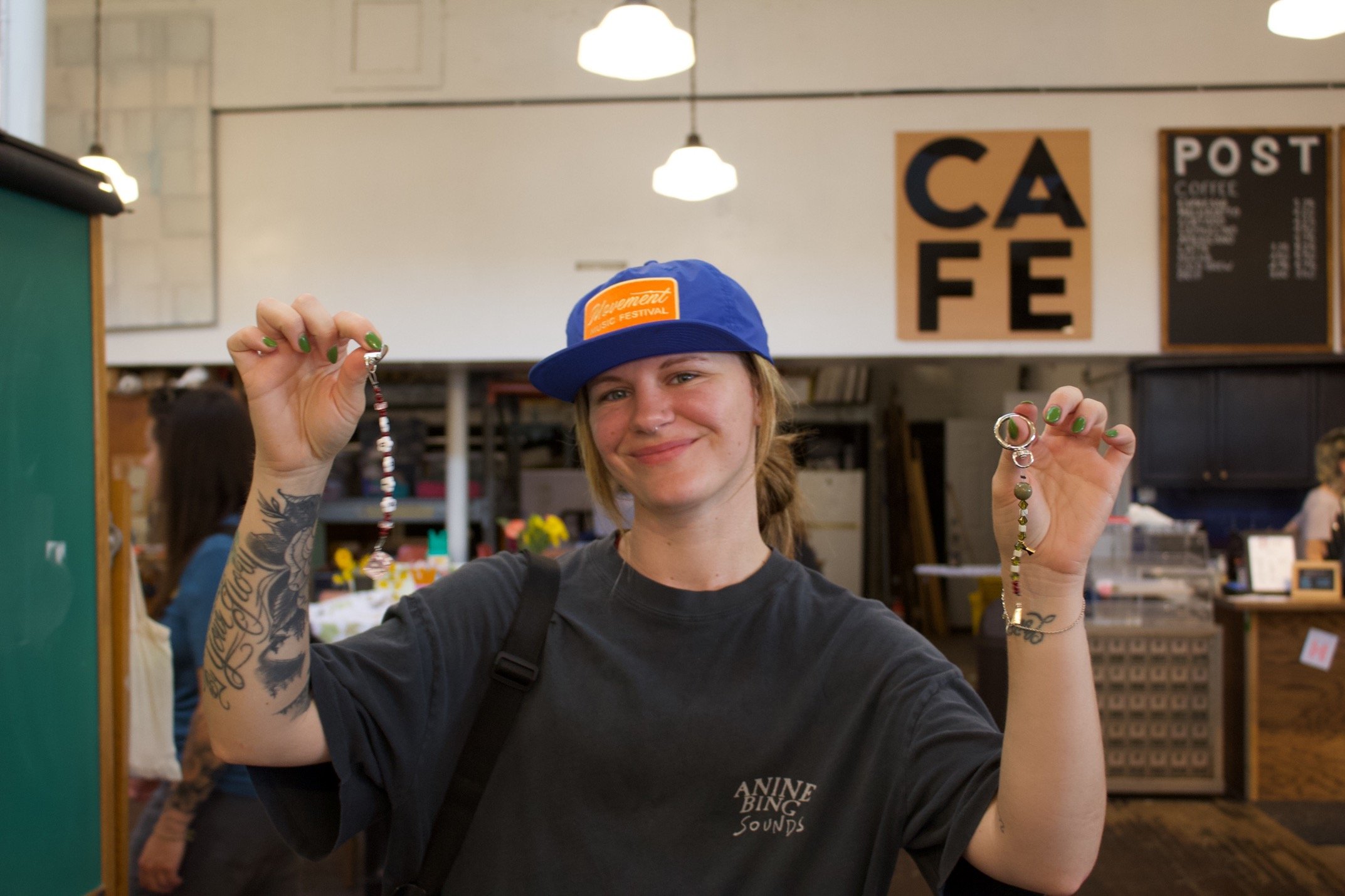 A smiling young woman with tattoos on her arms and green nail polish, wearing a blue cap and black t-shirt, holding handmade beaded jewelry in a marketplace.