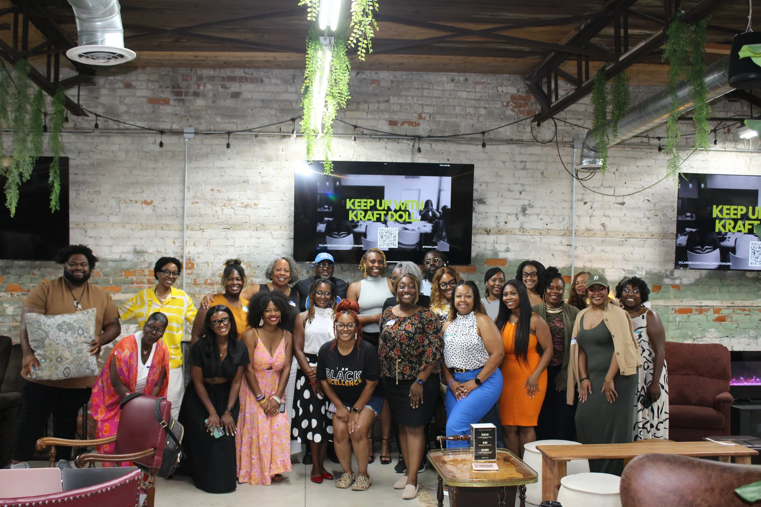 A group of women and men posing together indoors in a casual, artistic space with exposed brick walls, hanging greenery, and twin large screens displaying a message about Kraft Doli.