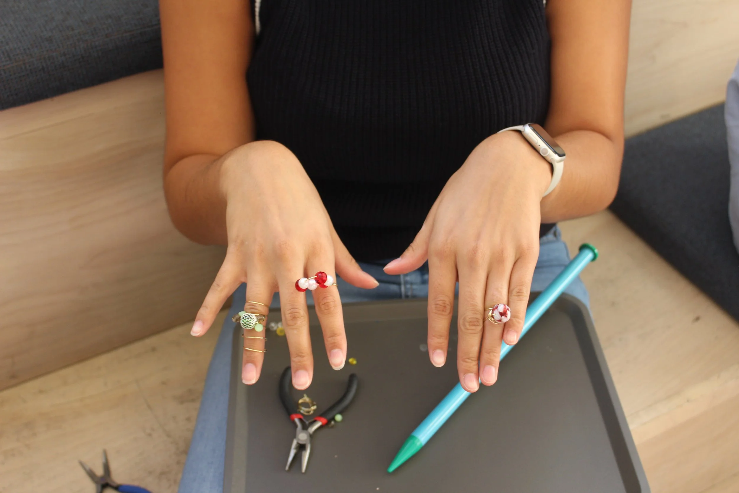 A person with several rings showing on their fingers, wearing a black sleeveless top, a white smartwatch, and a bracelet, displaying rings on their fingers, with jewelry and tools like pliers and a green pen on a black surface, possibly in a jewelry-making setting.