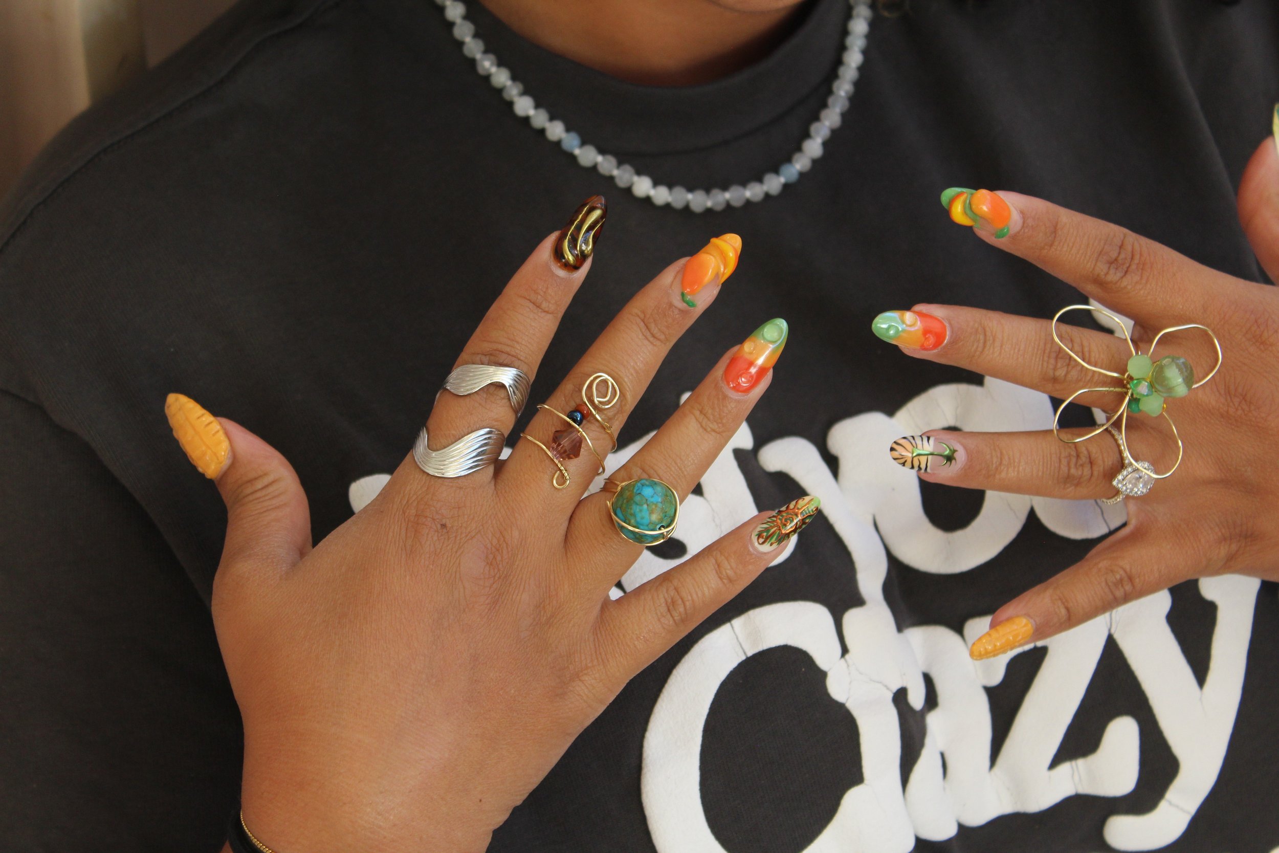 A person with manicured nails in colorful designs, wearing multiple rings on both hands, including a large circular turquoise ring, a gold butterfly ring, and a silver wavy band. The person is also wearing a beaded necklace and a black T-shirt.