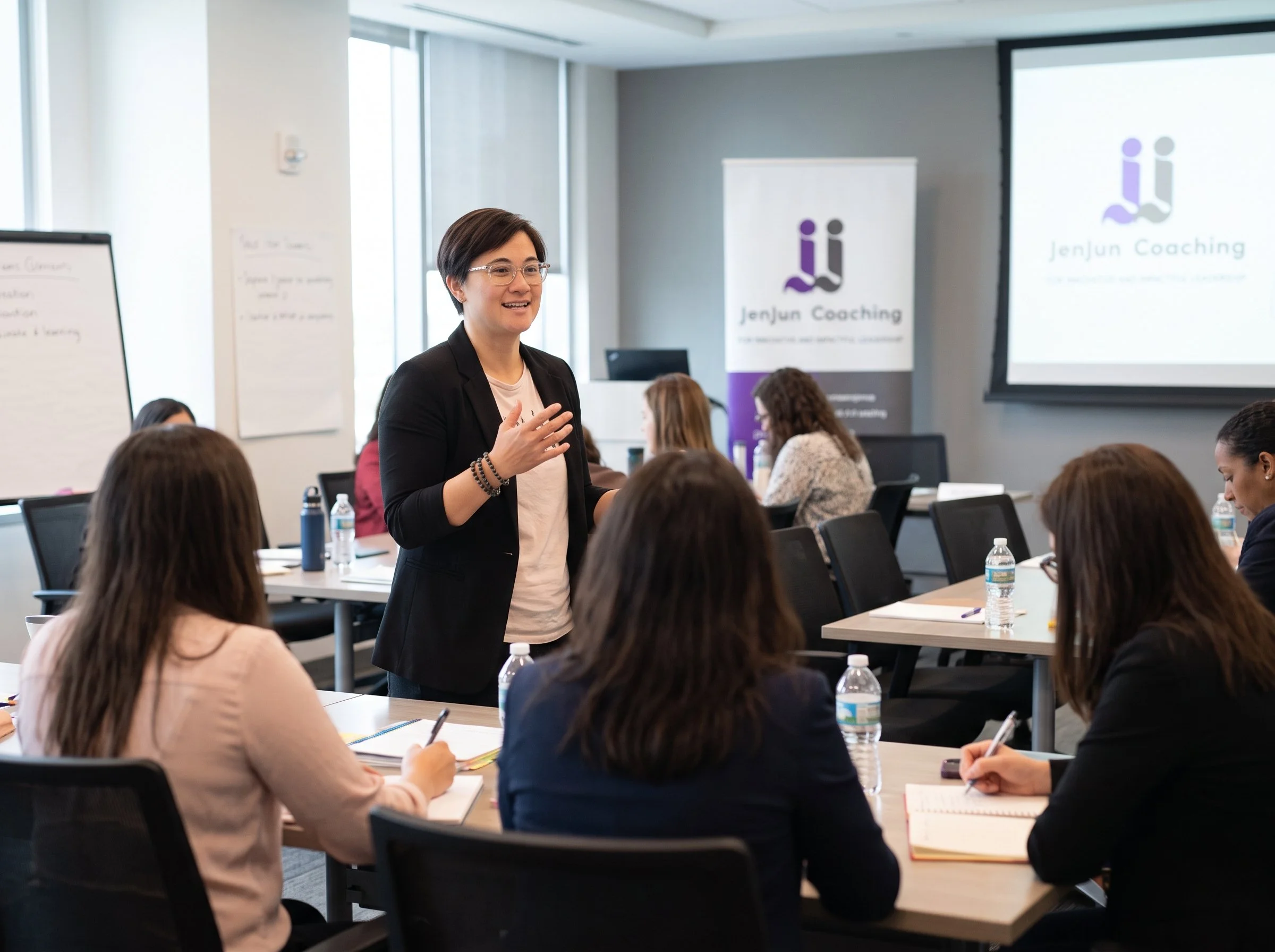 A woman giving a presentation in a conference room with a projector screen and a banner that reads 'Jenjun Coaching,' while several people seated at tables listen and take notes.