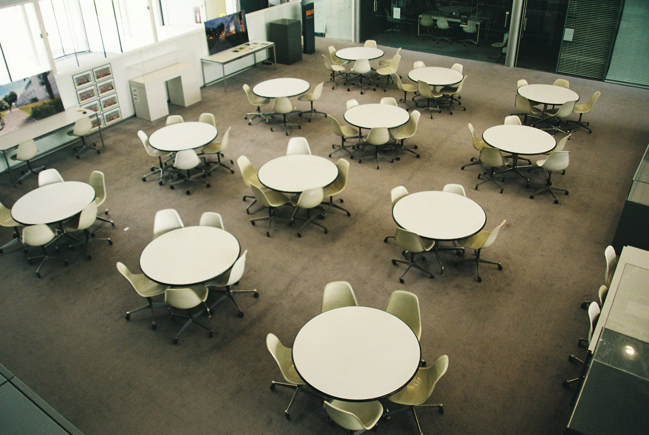 An empty modern conference room with round white tables surrounded by beige chairs, large windows on one side, and a display area with framed pictures and a television on the wall.