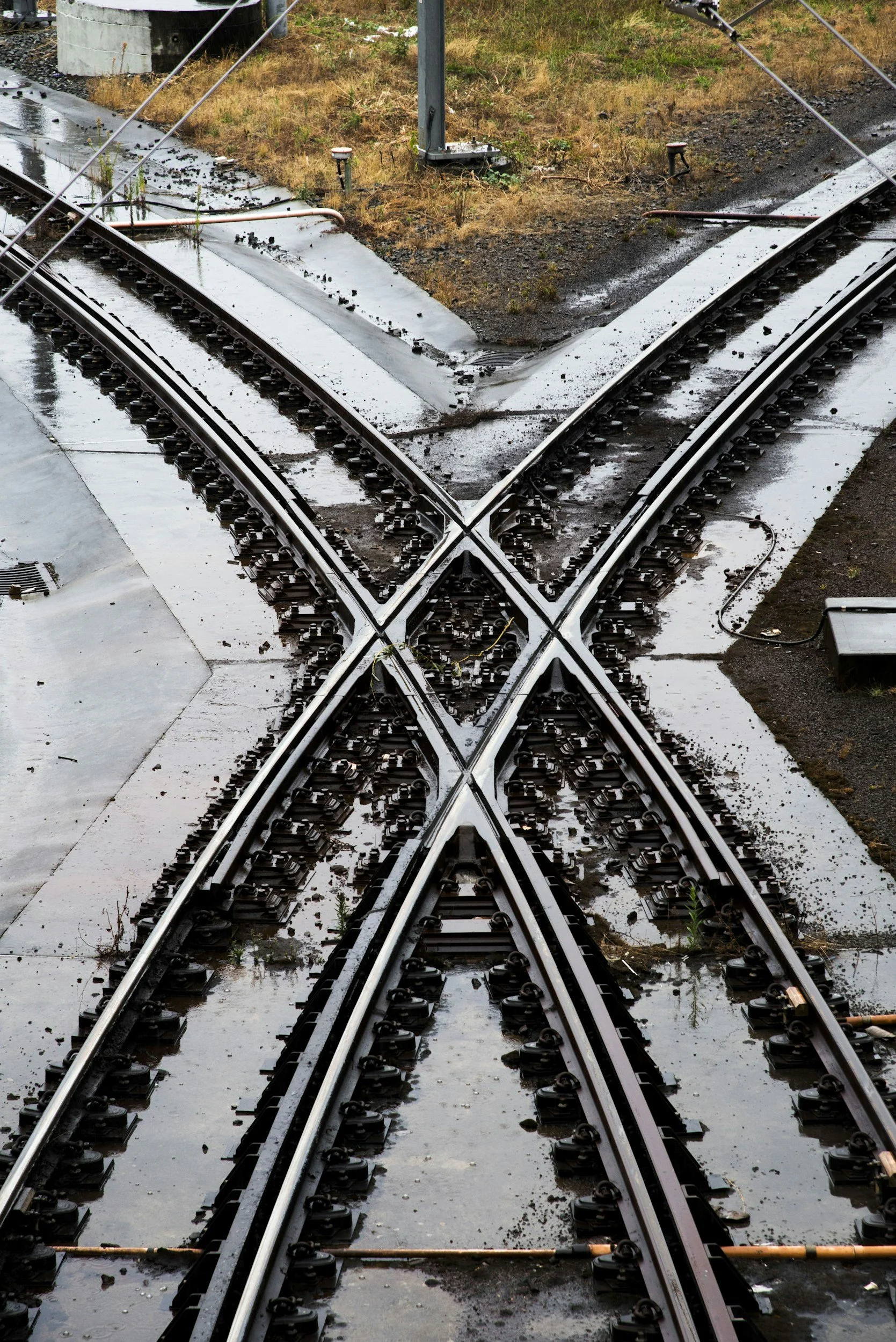 X-shaped railroad track switch with wet and muddy ground, grass, and some vegetation around the tracks.