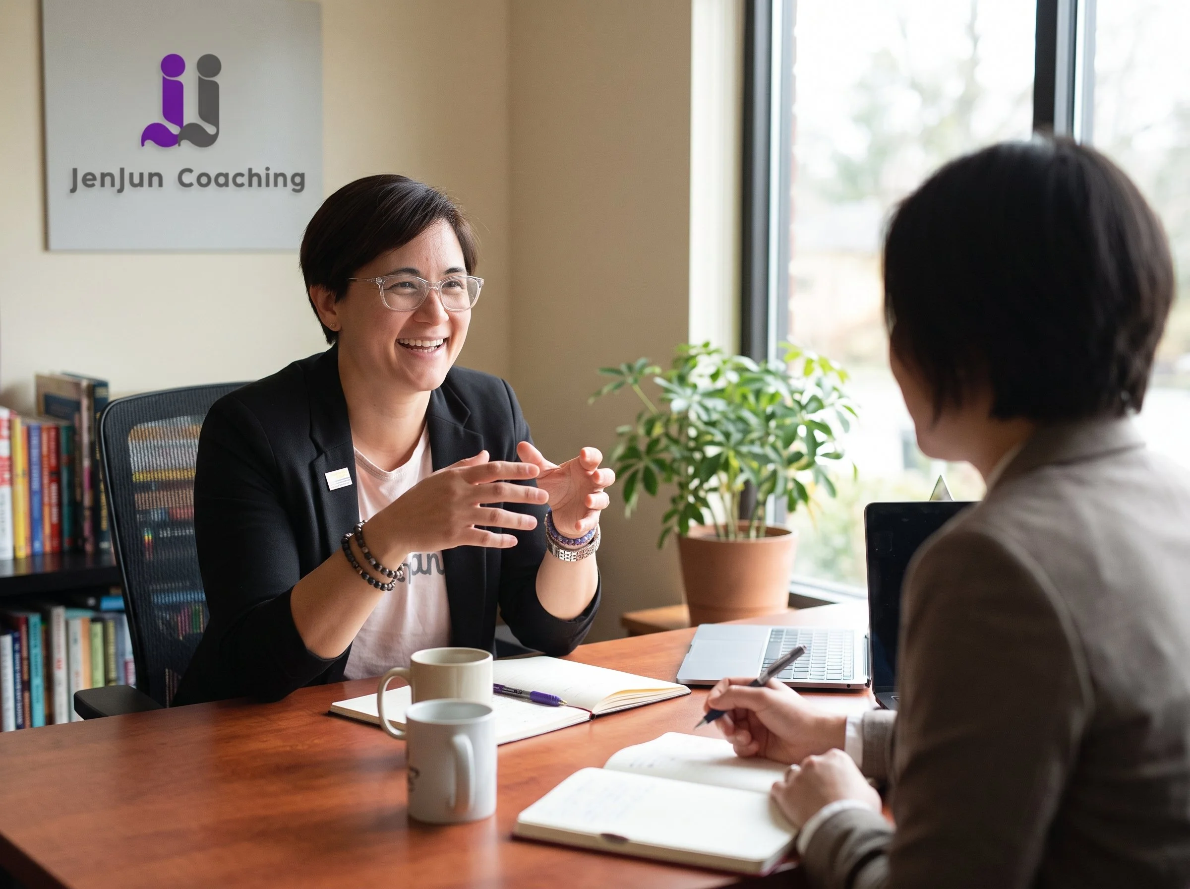 Two women are having a conversation in an office. One woman, with short hair and glasses, is smiling and gesturing with her hands. The other woman, seen from behind, is writing in a notebook. The office has a bookshelf, a potted plant, and a sign on the wall that reads "Jenjun Coaching." There are two coffee mugs and a laptop on the wooden table.