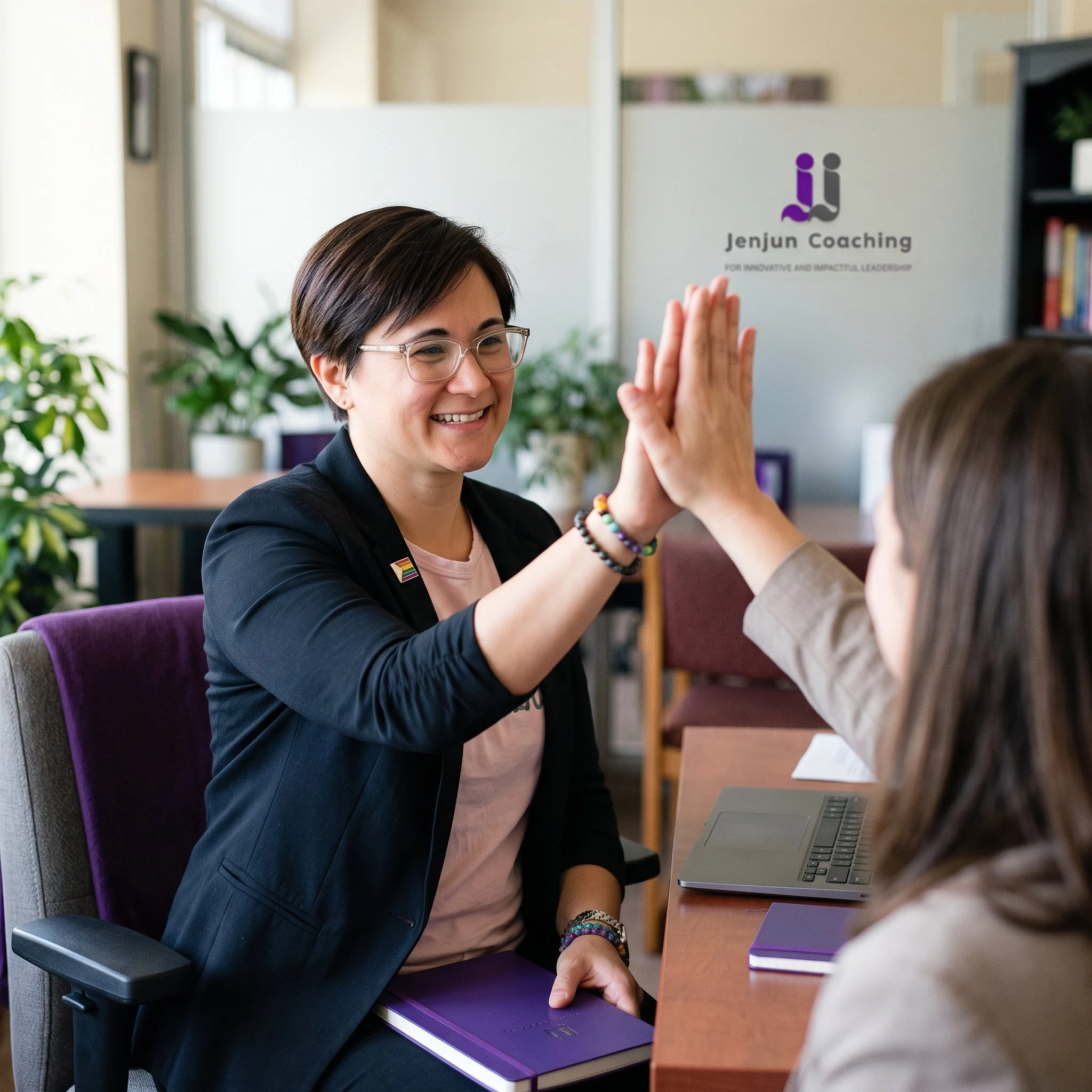 A woman with short dark hair, glasses, and a rainbow pin on her blazer is smiling and giving a high five to another person in an office setting. There is a laptop, colorful notebooks, and green plants in the background.