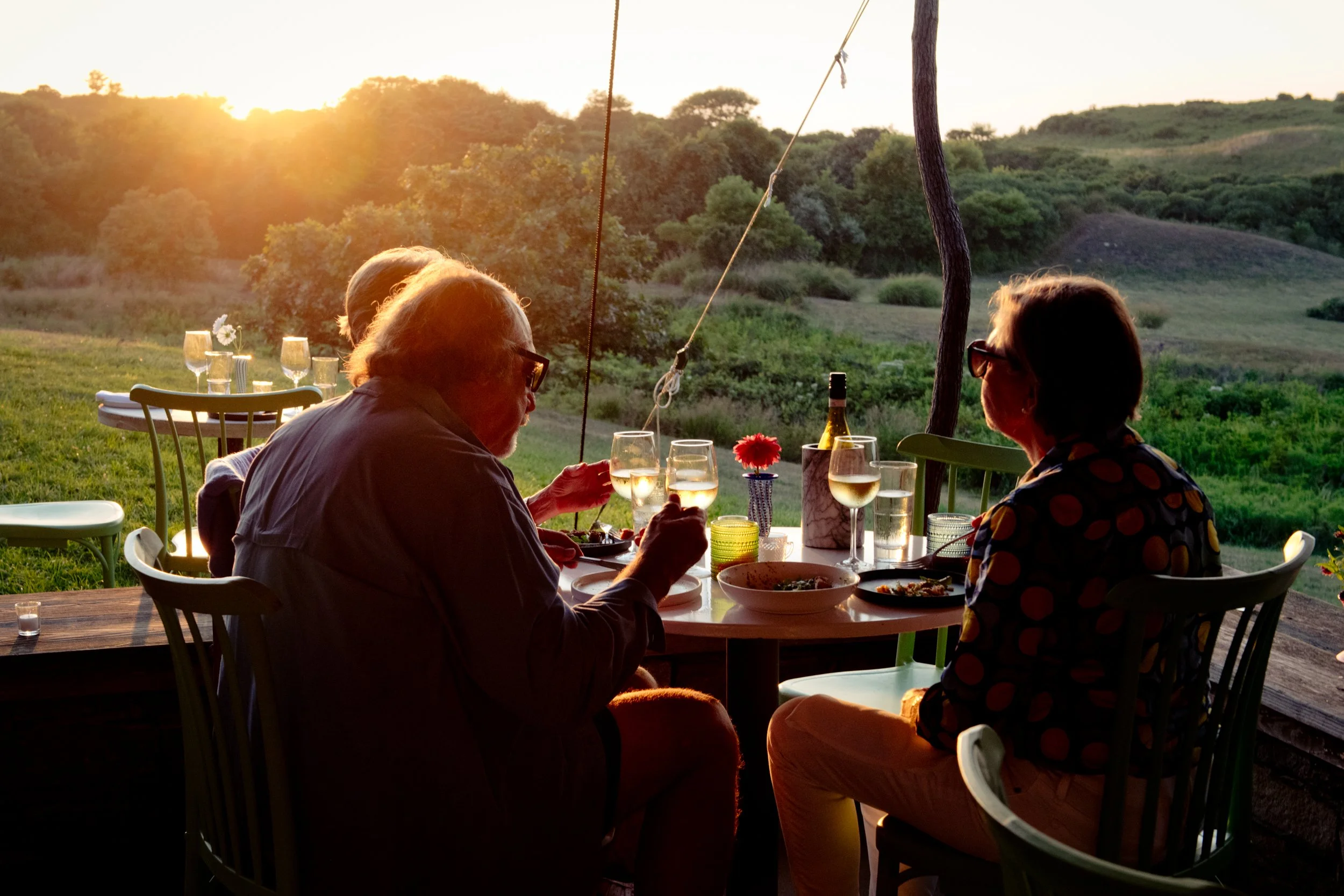 People dining outdoors at sunset in a rural landscape, with glasses of white wine, food on the table, and green hills in the background.
