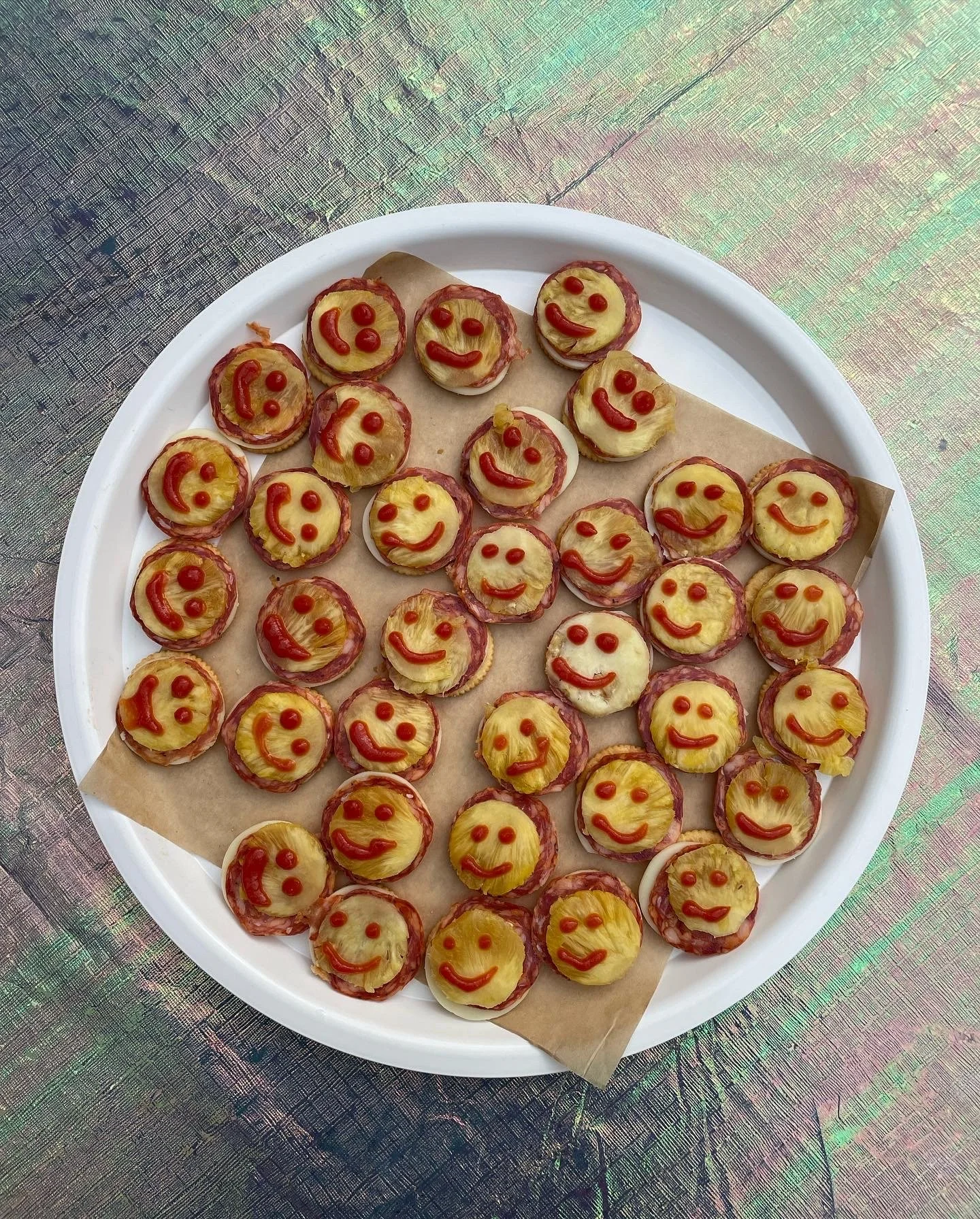 Miniature pizza snacks topped with pineapple and ketchup faces arranged on a white tray with parchment paper, on a holographic surface.