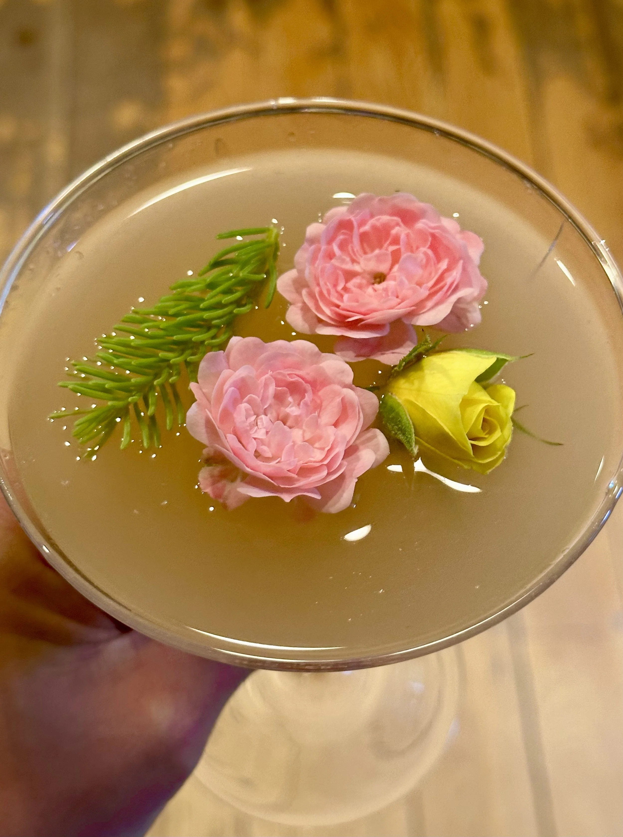 A cocktail glass containing a clear drink with floating pink roses, a yellow rosebud, and a green pine needle branch.