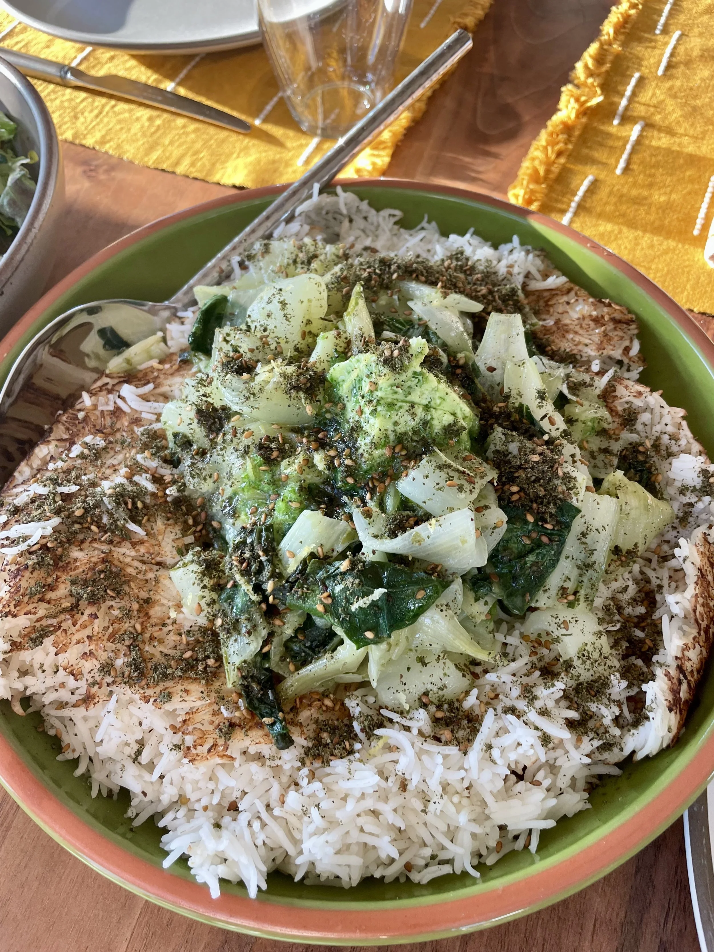 A bowl of white rice topped with cooked greens, chopped vegetables, and sprinkled with seasonings and spices.