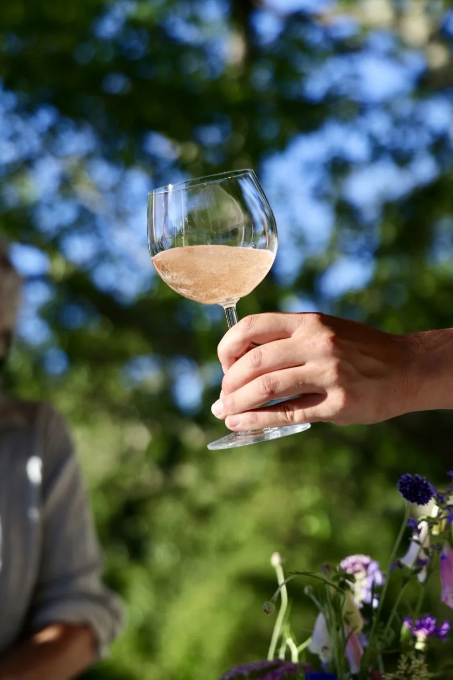 A hand holding a glass of rose wine outdoors with trees and blue sky in the background at a garden party in Chilmark