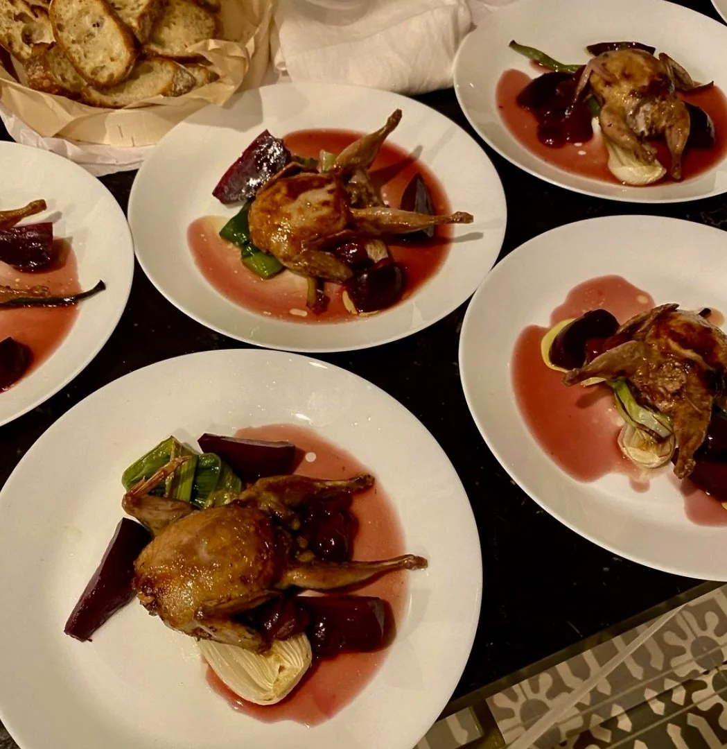 Multiple plates with roasted quail, vegetables, and sauce, along with a basket of bread.