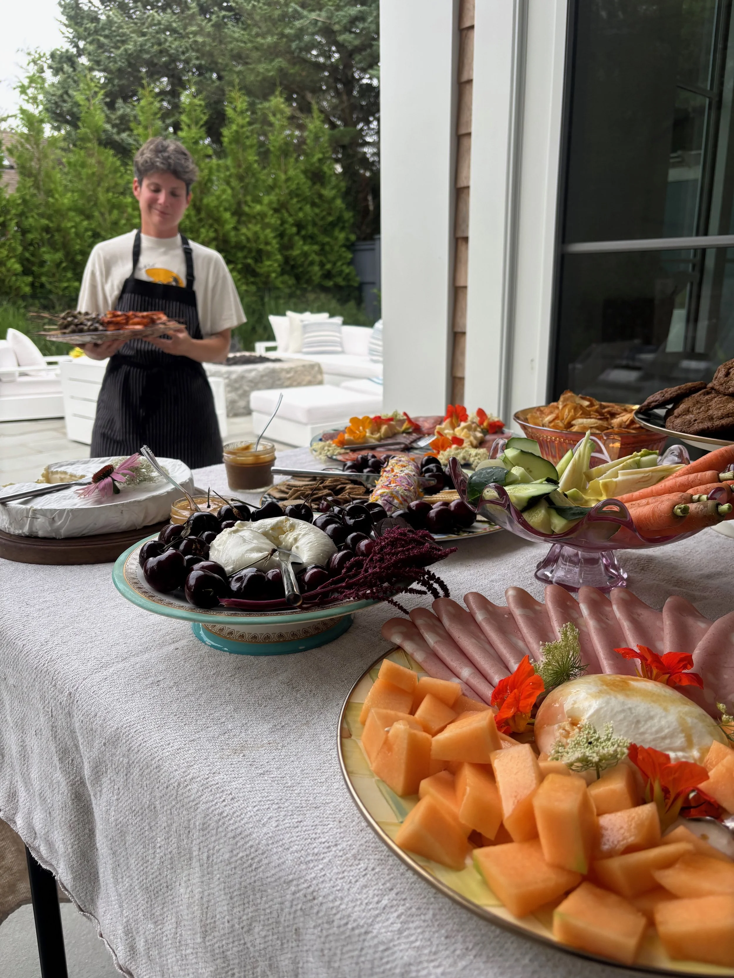 A woman holding a tray of grilled food near a table of assorted cheeses, fruits, vegetables, and meats set up for a buffet outside, with greenery and outdoor furniture in the background.
