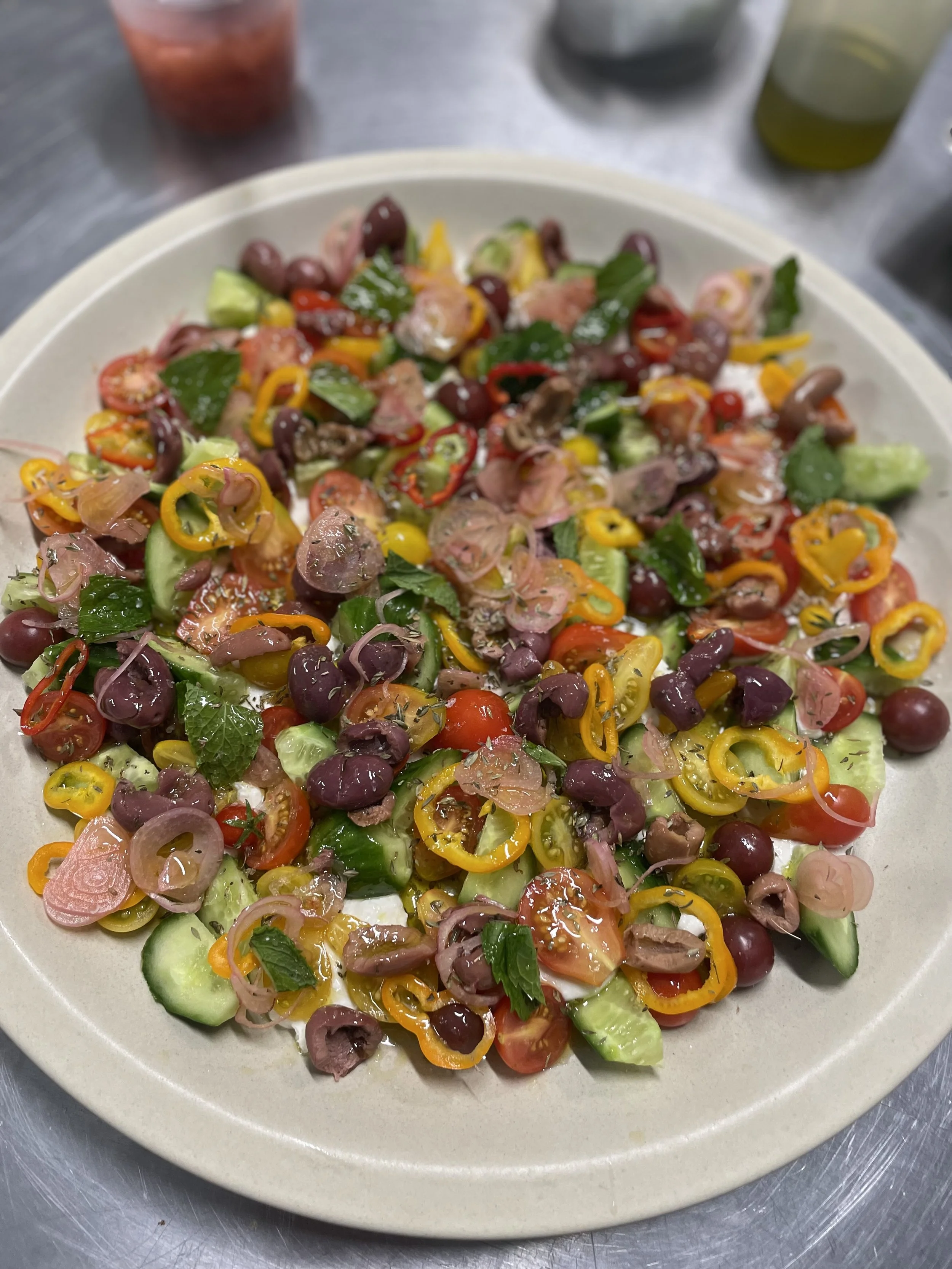 A plate of a colorful salad with cherry tomatoes, cucumbers, yellow bell peppers, red onions, black olives, and fresh herbs.