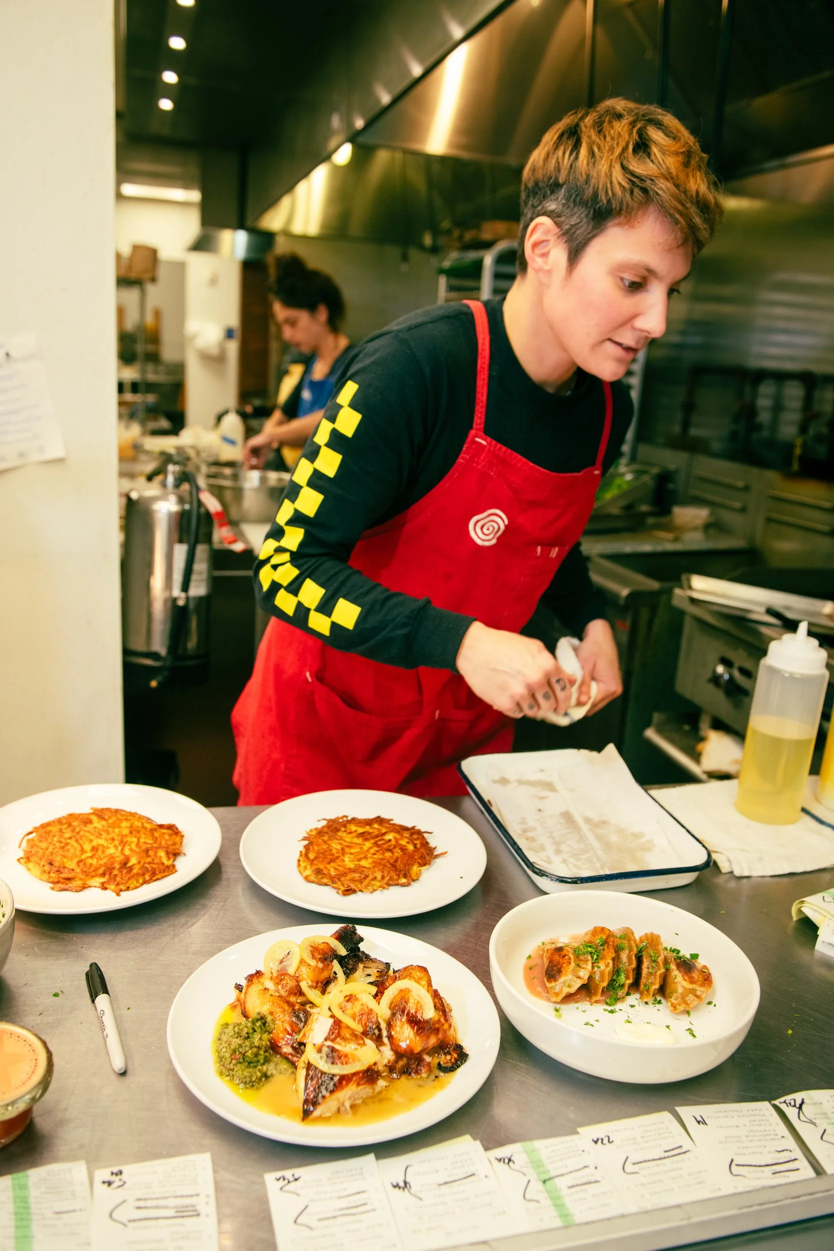 A woman with short hair wearing a black shirt and red apron preparing food in a commercial kitchen, with plates of cooked dishes including crispy fried items and garnished pieces on the counter.