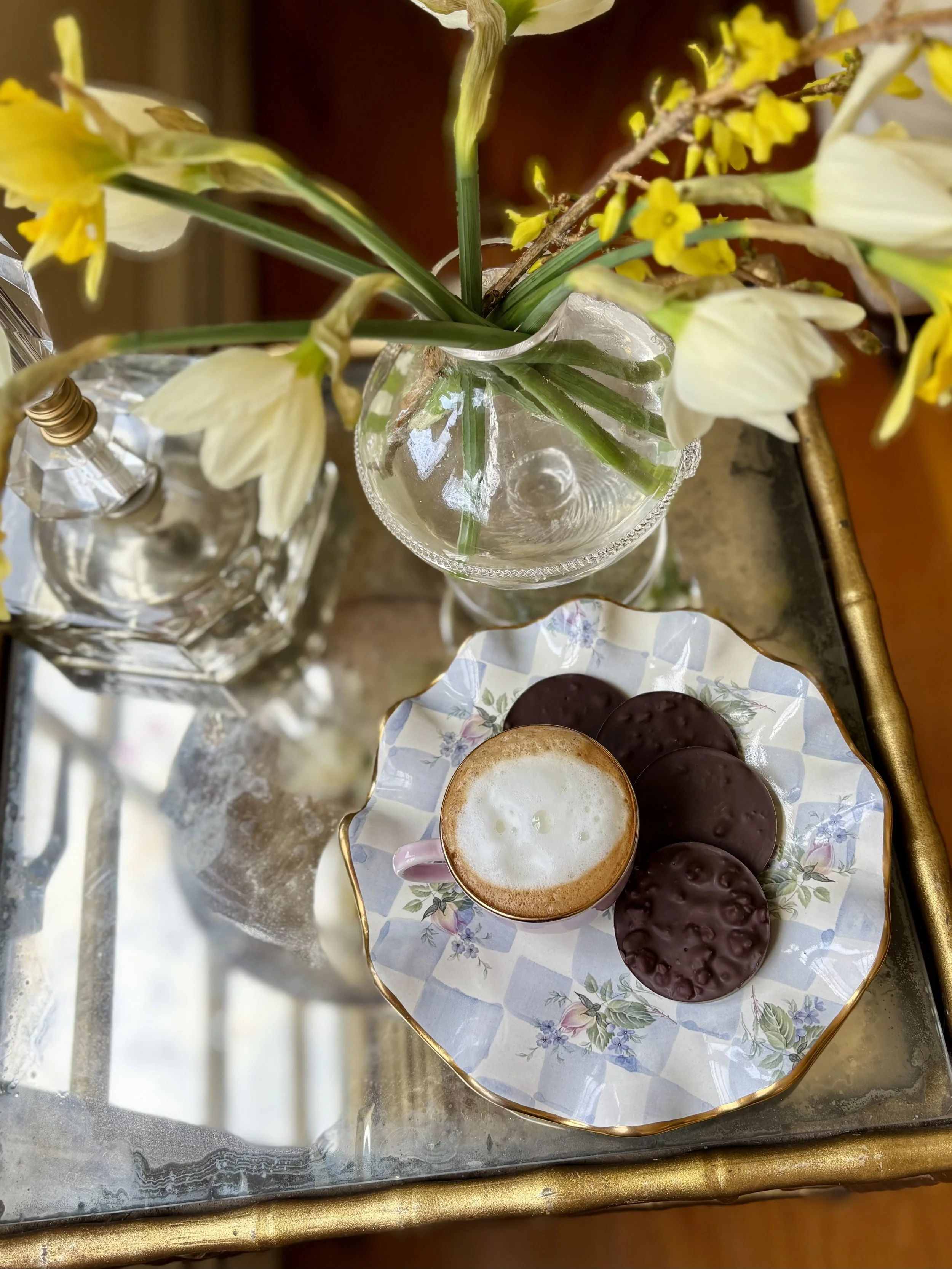 A glass vase with yellow and white flowers, a square glass perfume bottle, and a plate with a pink mug of frothy coffee and four chocolate covered cookies on a glass table with a gold edge.