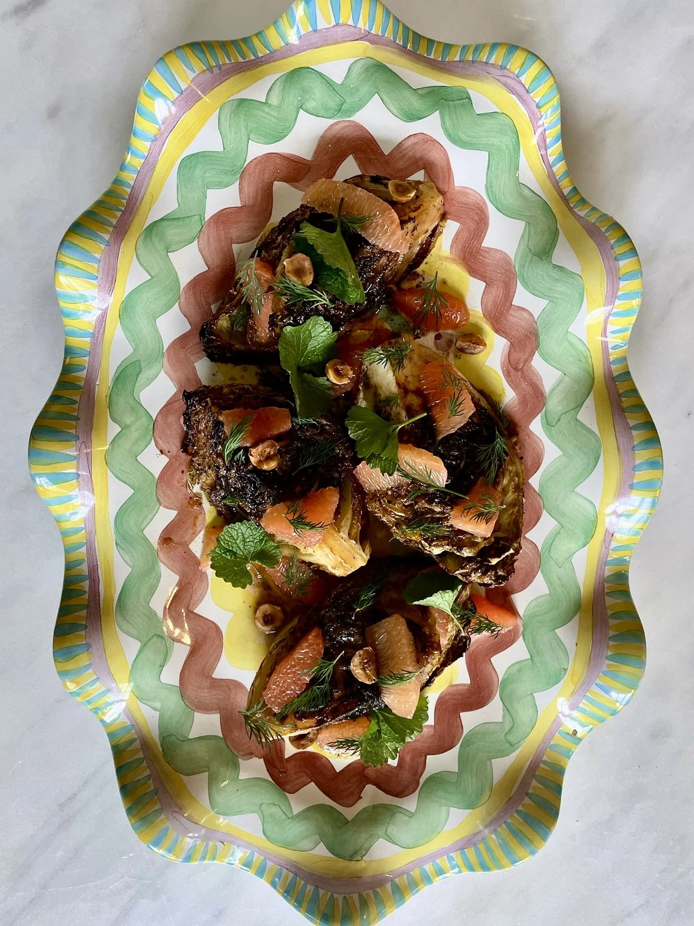 A decorative platter with roasted fish topped with herbs, citrus slices, and nuts, garnished with fresh herbs on a colorful, patterned ceramic dish.