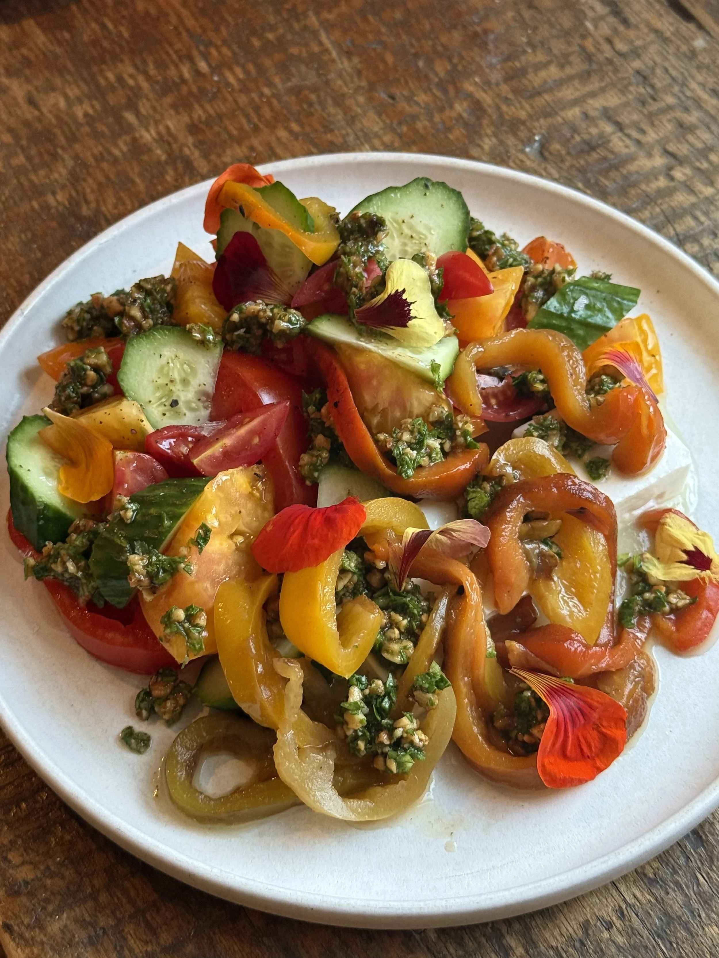 Colorful salad with sliced cucumbers, cherry tomatoes, yellow, red, and orange bell peppers, topped with edible flower petals, served on a white plate on a wooden table.