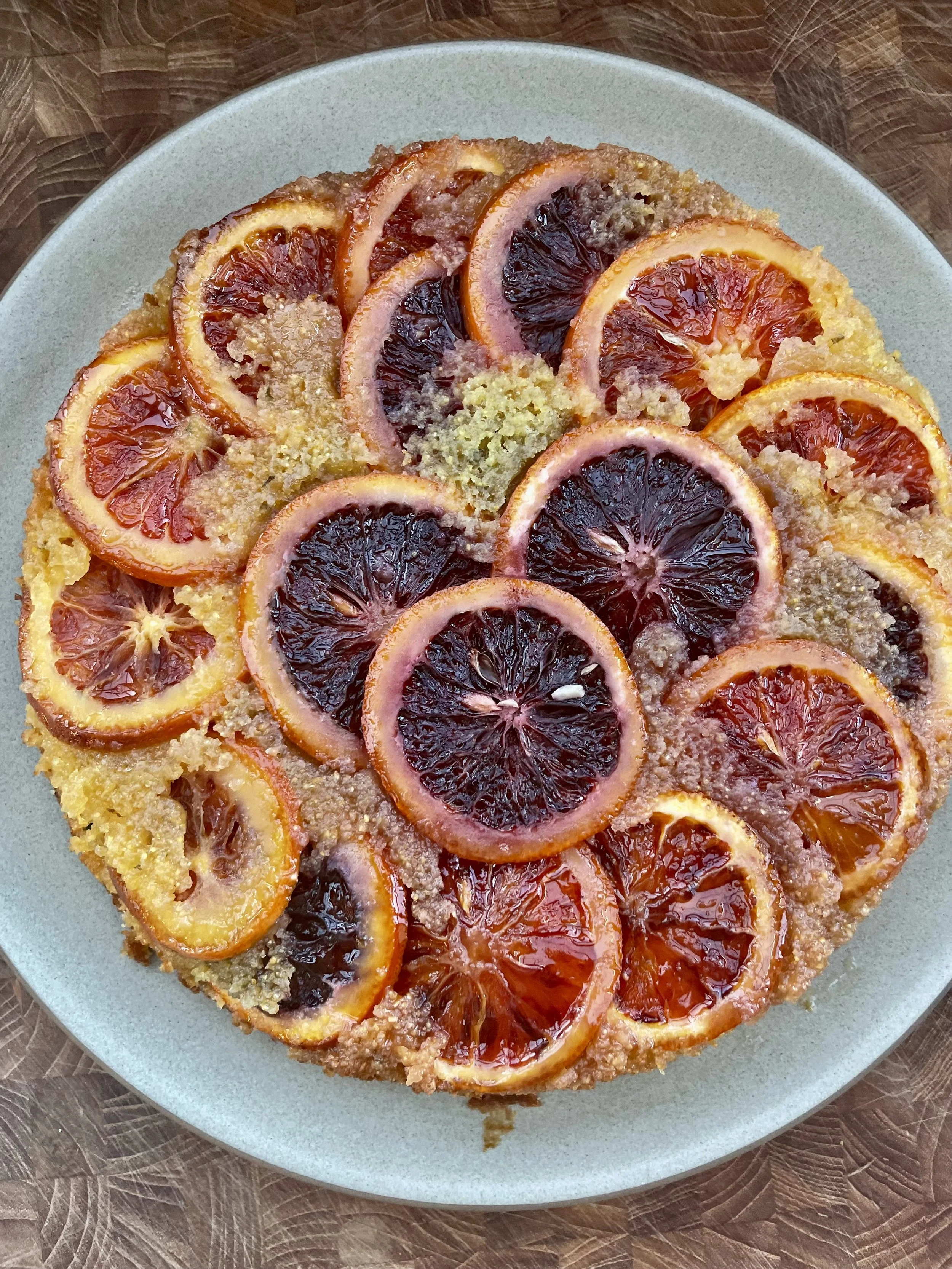 A round fruit tart topped with sliced blood oranges and regular oranges, garnished with sugar granules on a ceramic plate.