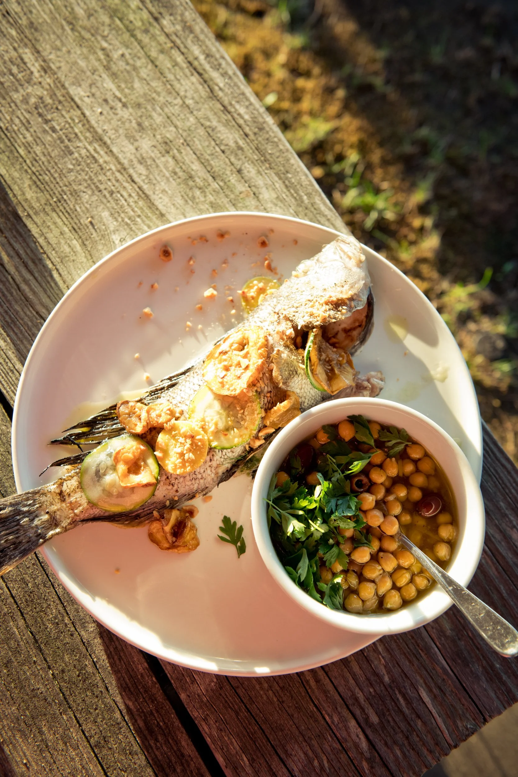 A plate with a cooked fish garnished with sliced vegetables and garlic, served with a side of chickpea soup garnished with herbs, on a rustic wooden surface outdoors.