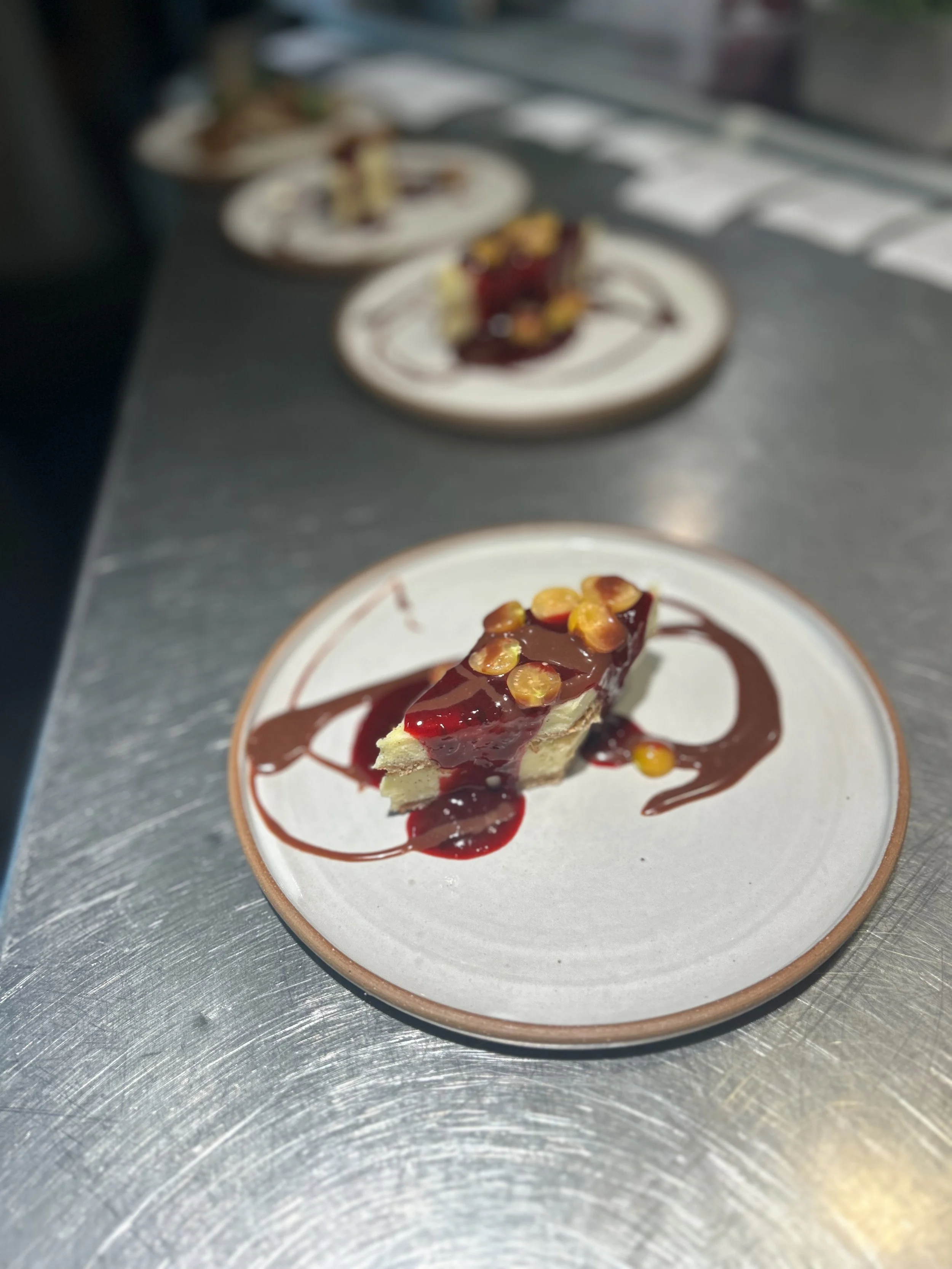 A slice of cheesecake with chocolate ganache and caramelized nuts on top, drizzled with chocolate and caramel sauces on a white plate.