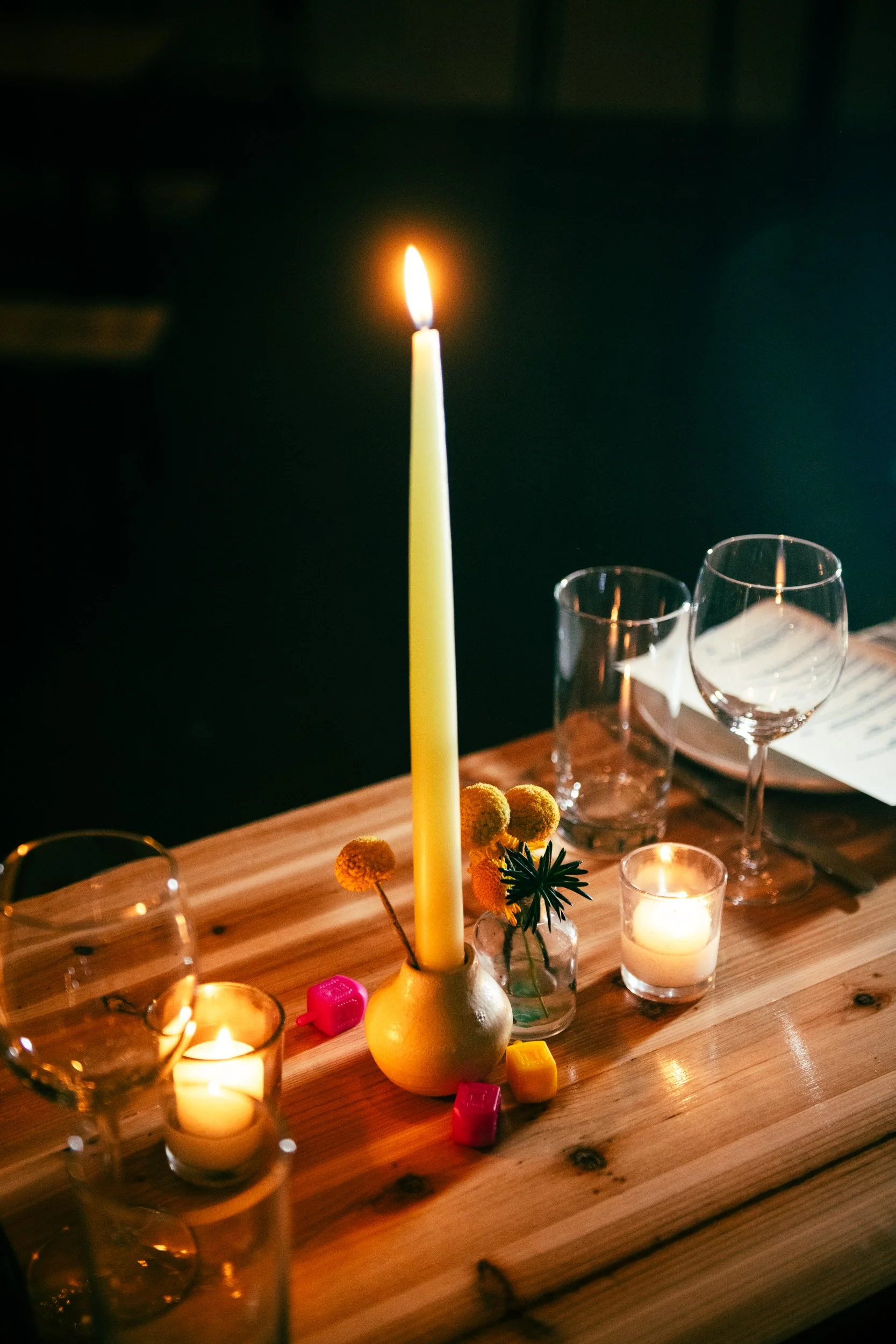 A lit yellow candle in a small vase with yellow flowers on a wooden table, surrounded by candles, glasses, and a menu, in a dimly lit setting.