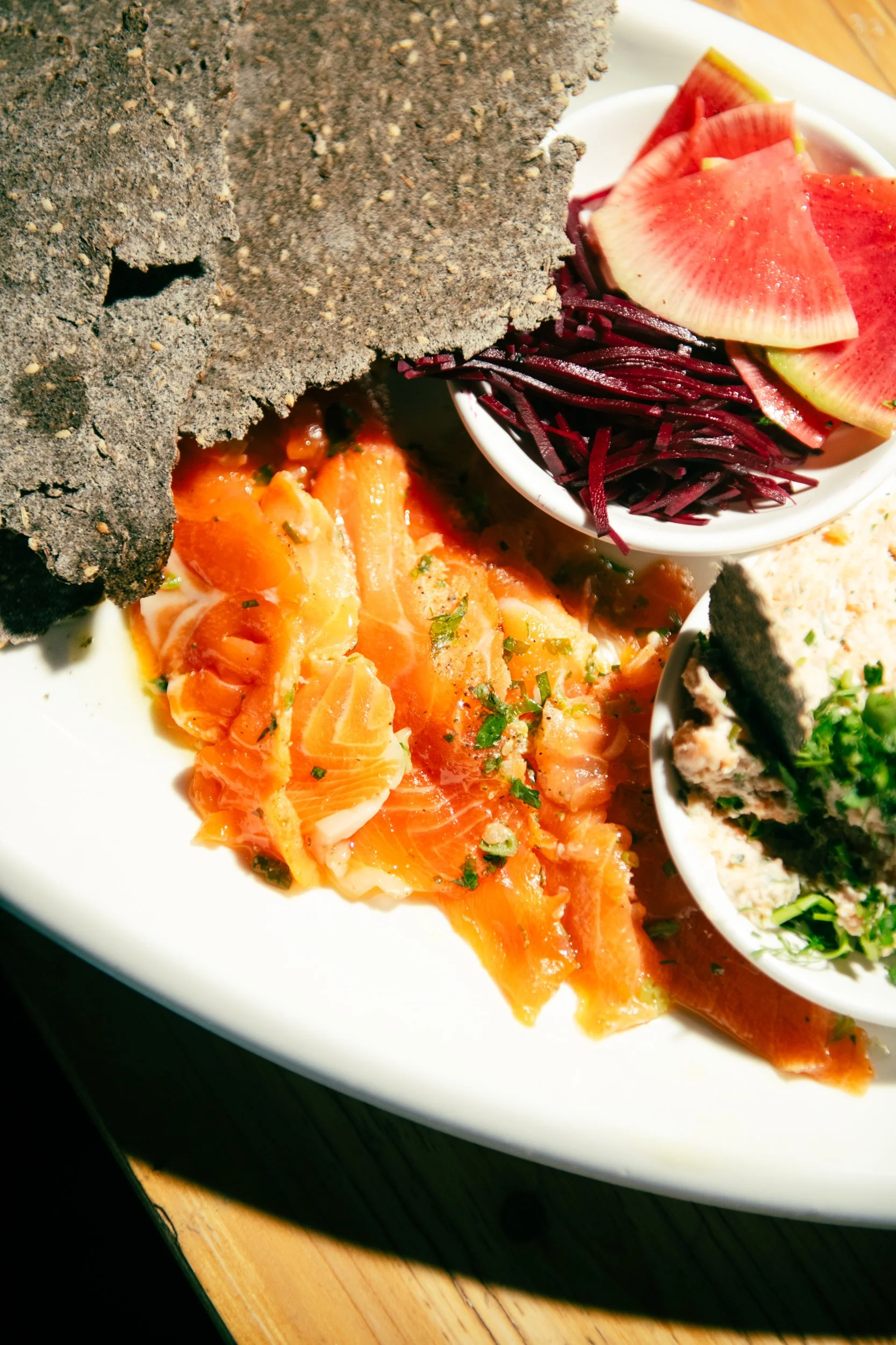A white plate with smoked salmon, fresh vegetables, and black charcoal bread.