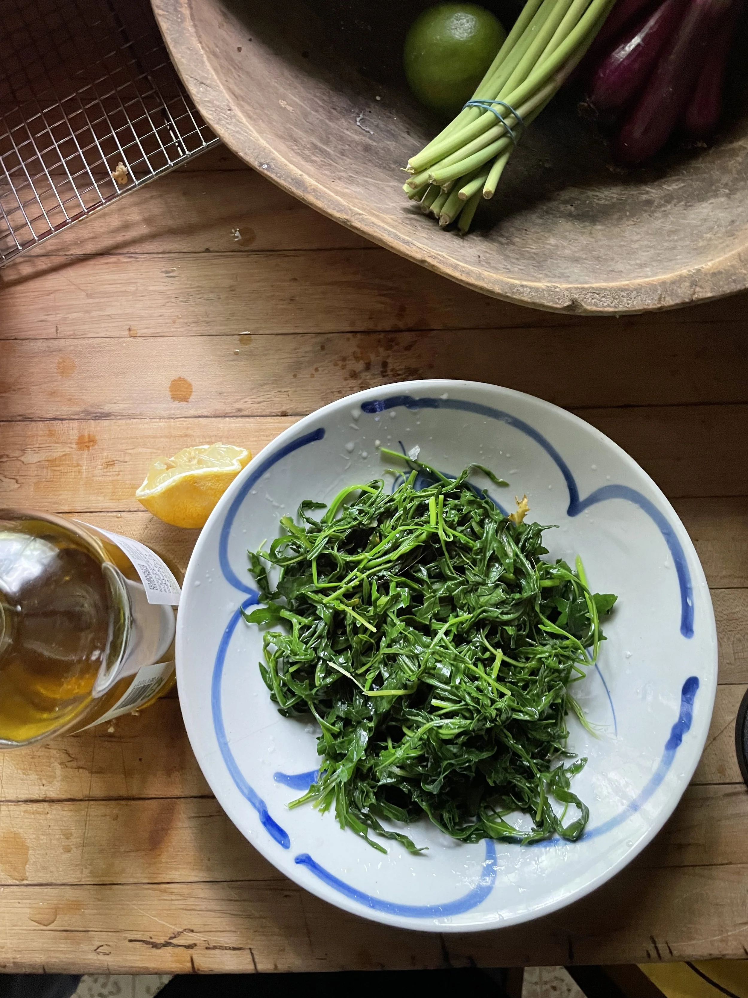A white plate with a blue border filled with fresh green arugula leaves on a wooden table, with a lemon wedge and a partially visible bottle of beer nearby, and a wooden bowl containing limes, green onions, and purple onions in the background.