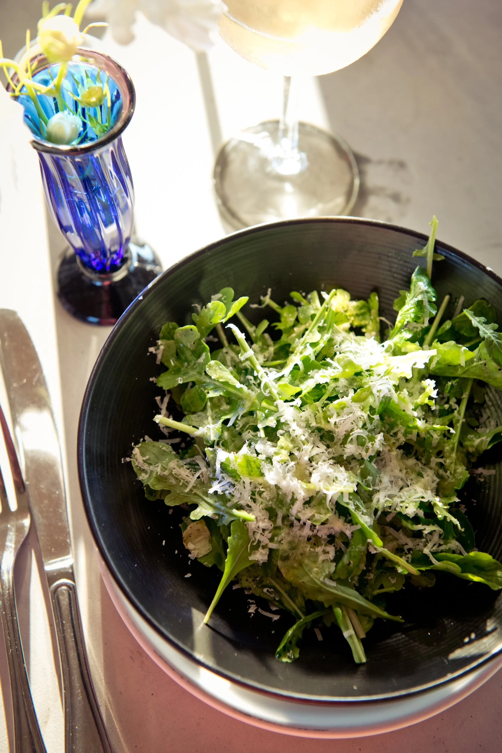 Fresh green salad with shredded cheese in a black bowl, glass of white wine, and a blue vase with yellow flowers on a table.