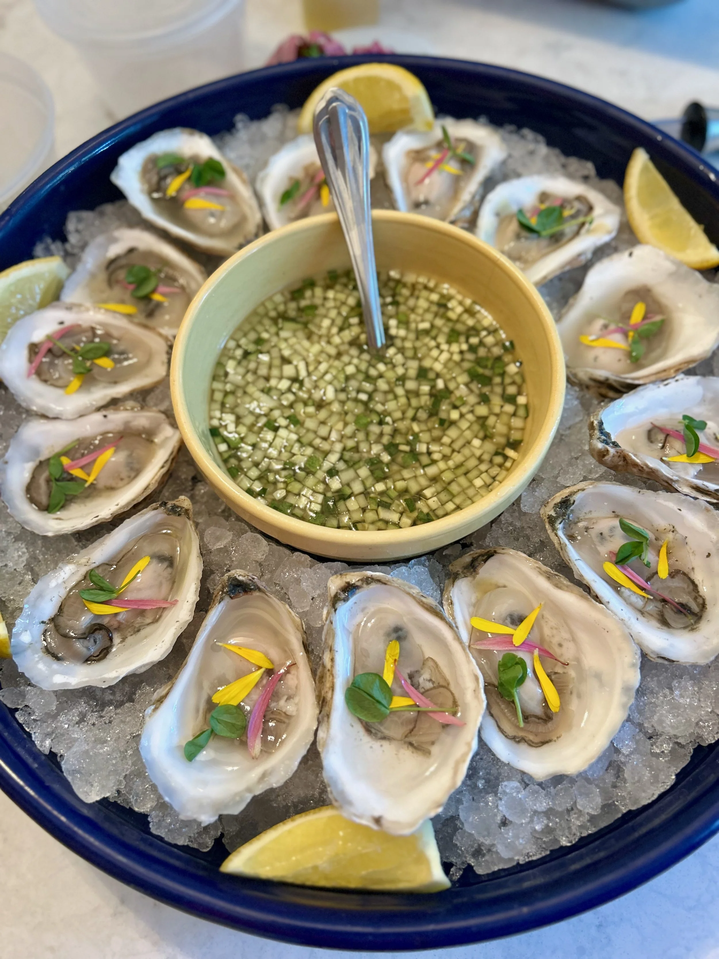 A platter of fresh oysters on ice garnished with lemon wedges, small herbs, and colorful edible flowers, served with a dipping sauce containing diced cucumber or similar vegetable.