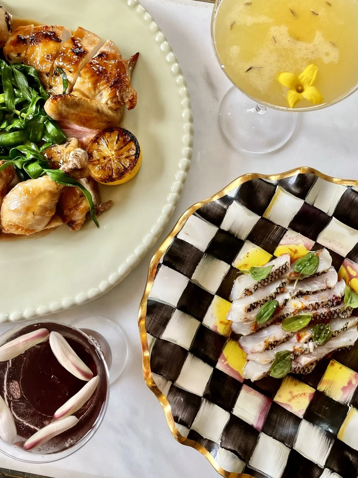 A meal with grilled chicken, sautéed greens, roasted lemon, sliced sashimi, a glass of a yellow cocktail with yellow flowers, and a glass of a dark beverage topped with flower petals.