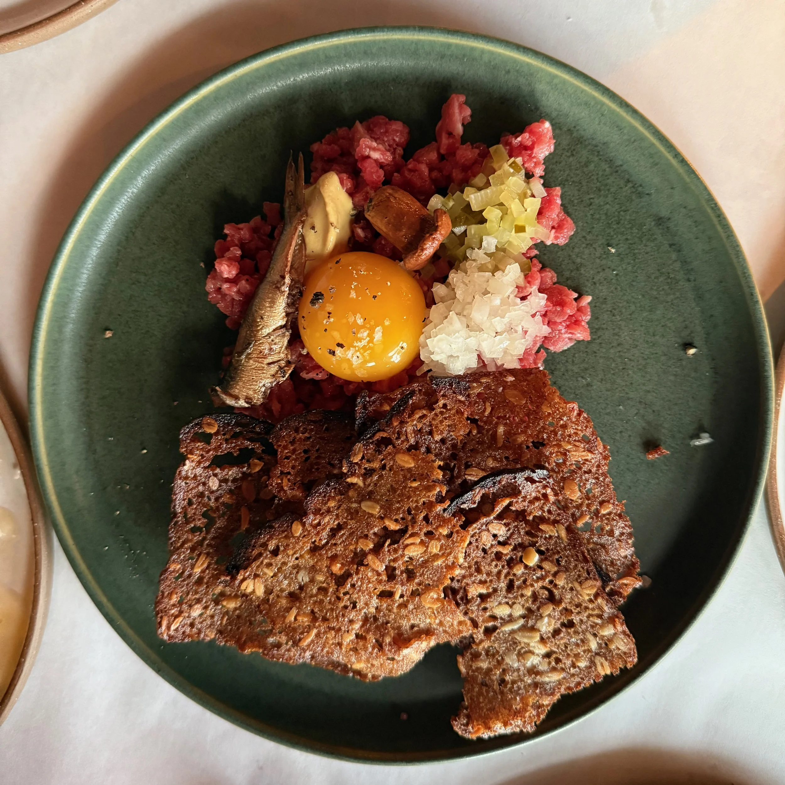 A green ceramic plate with a cooked grain and seed bread, a raw egg yolk, a small grilled fish, chopped onion, and diced celery on a white surface.