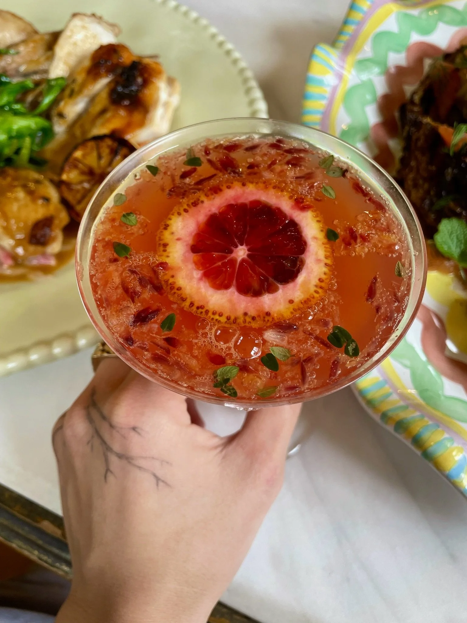 A hand holding a pink cocktail with an ice cube and a slice of blood orange inside. Plates of food are visible in the background.