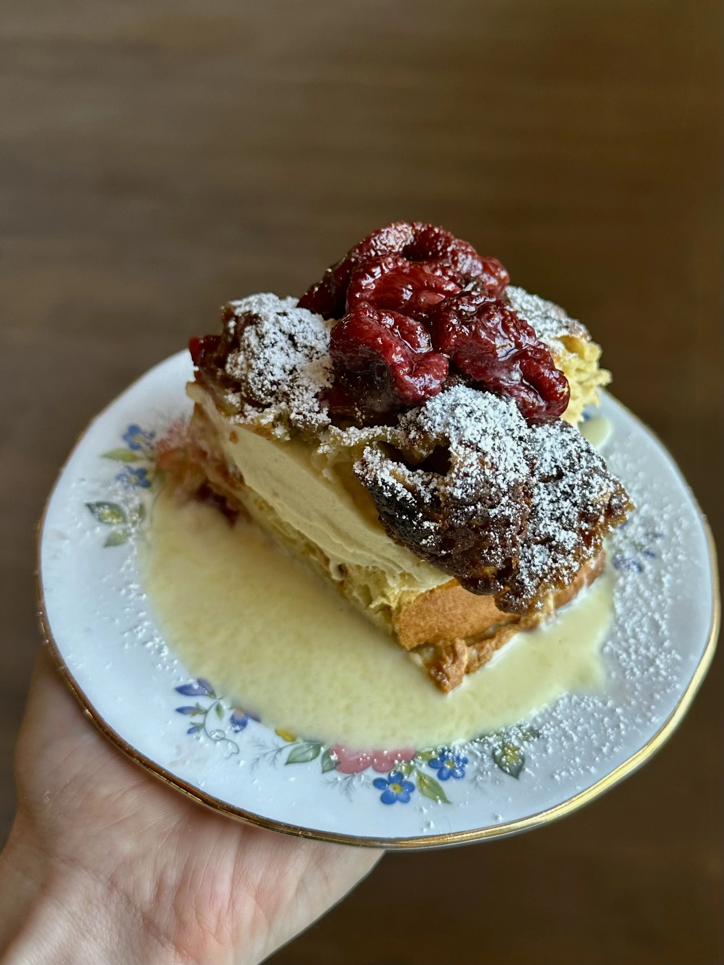 A dessert slice with raspberries, powdered sugar, and sauce on a floral-patterned plate.