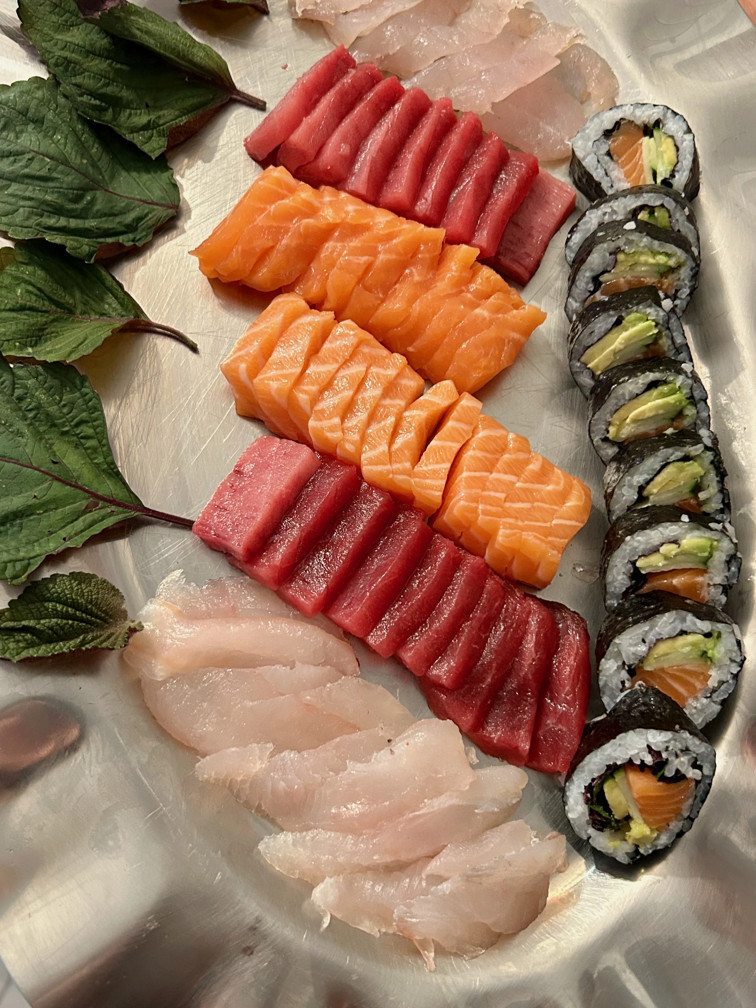 Assorted sushi and sashimi pieces arranged on a metal tray with green leaves for decoration.