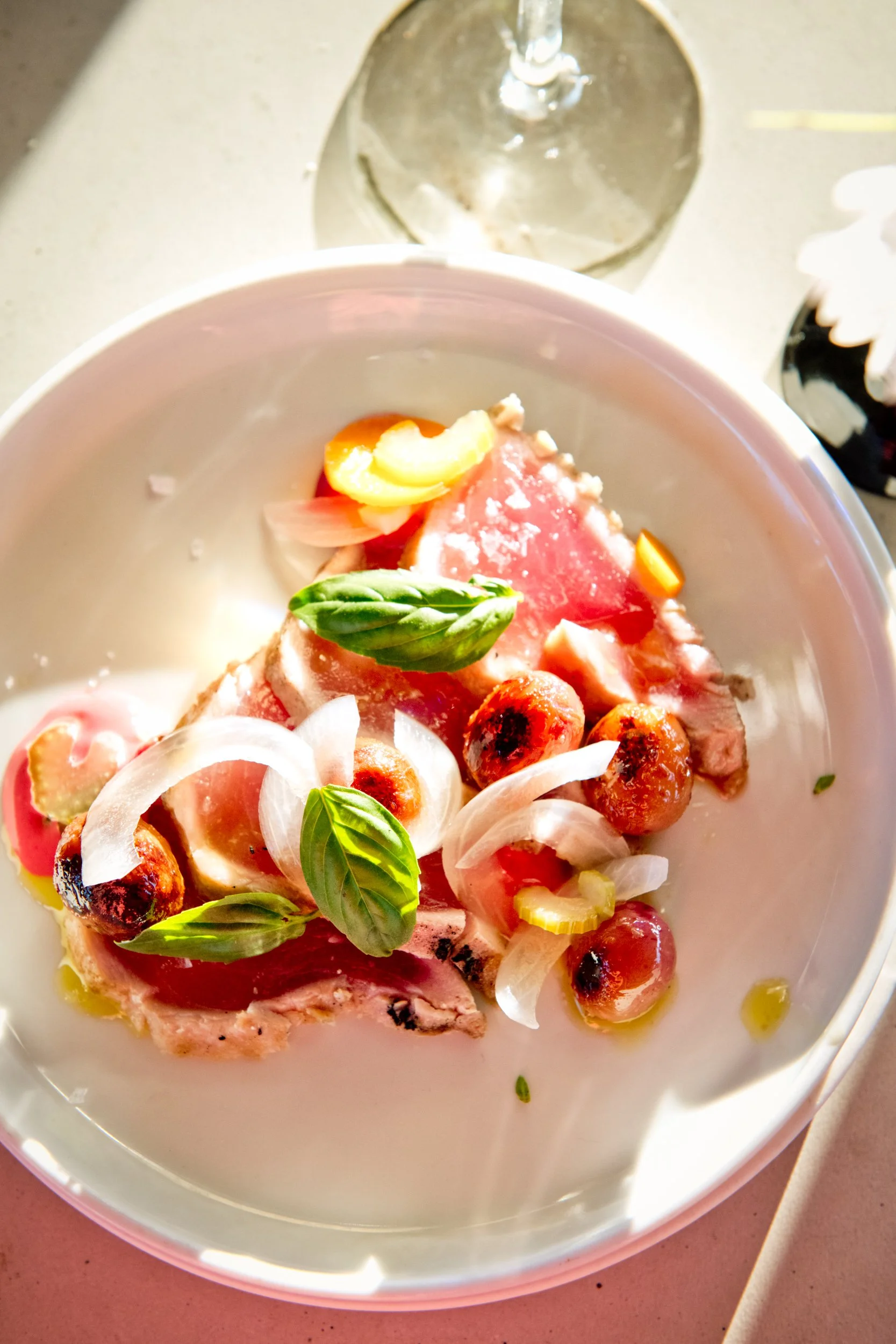 A plate of sliced cooked meat garnished with fresh basil leaves, roasted cherry tomatoes, sliced onions, and small yellow peppers, with a glass of white wine in the background.