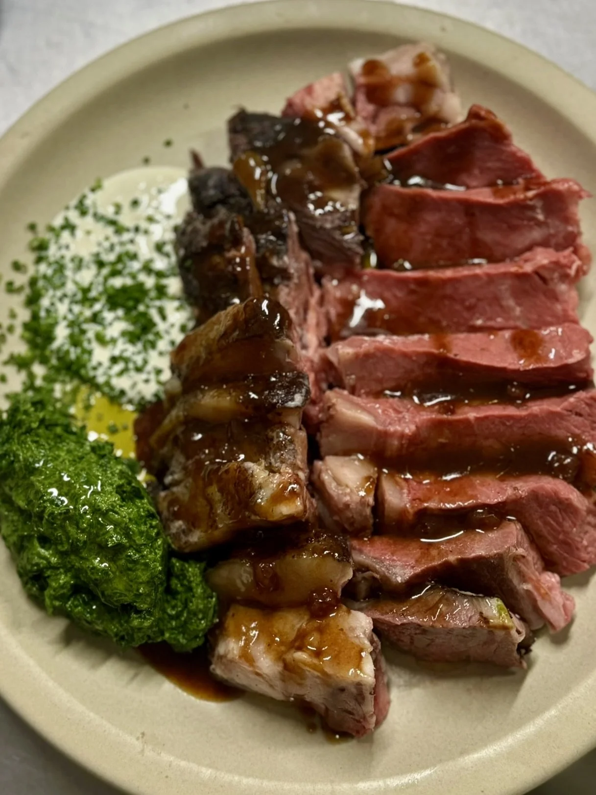 Sliced medium-rare steak with brown gravy, a serving of green pesto, and chopped chives on a beige plate.