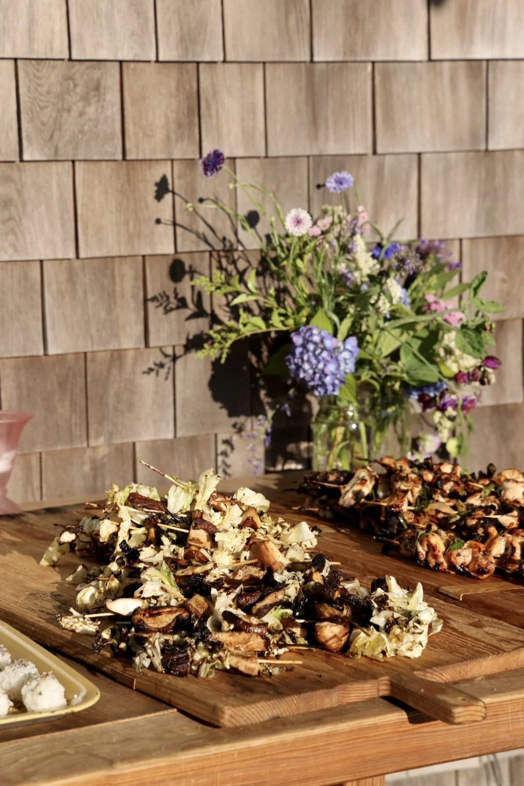 Wooden table with grilled chicken skewers and chopped vegetables in the foreground, and a bouquet of purple, pink, white and blue flowers in a jar against a wooden shingle wall in the background in Chilmark