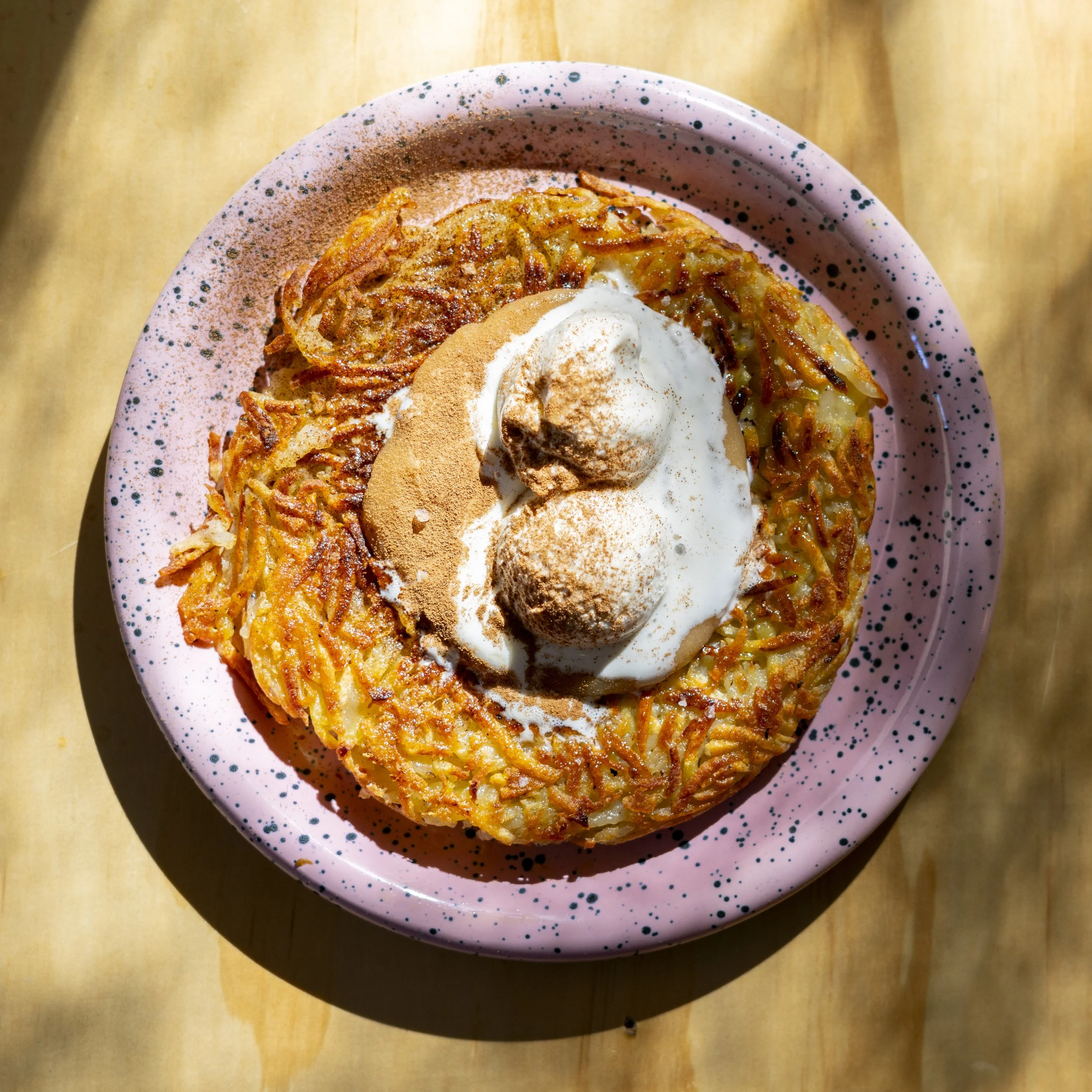 A plate of crispy hash browns topped with whipped cream, cinnamon, and two scoops of ice cream or similar dessert, on a pink speckled plate on a wooden surface.