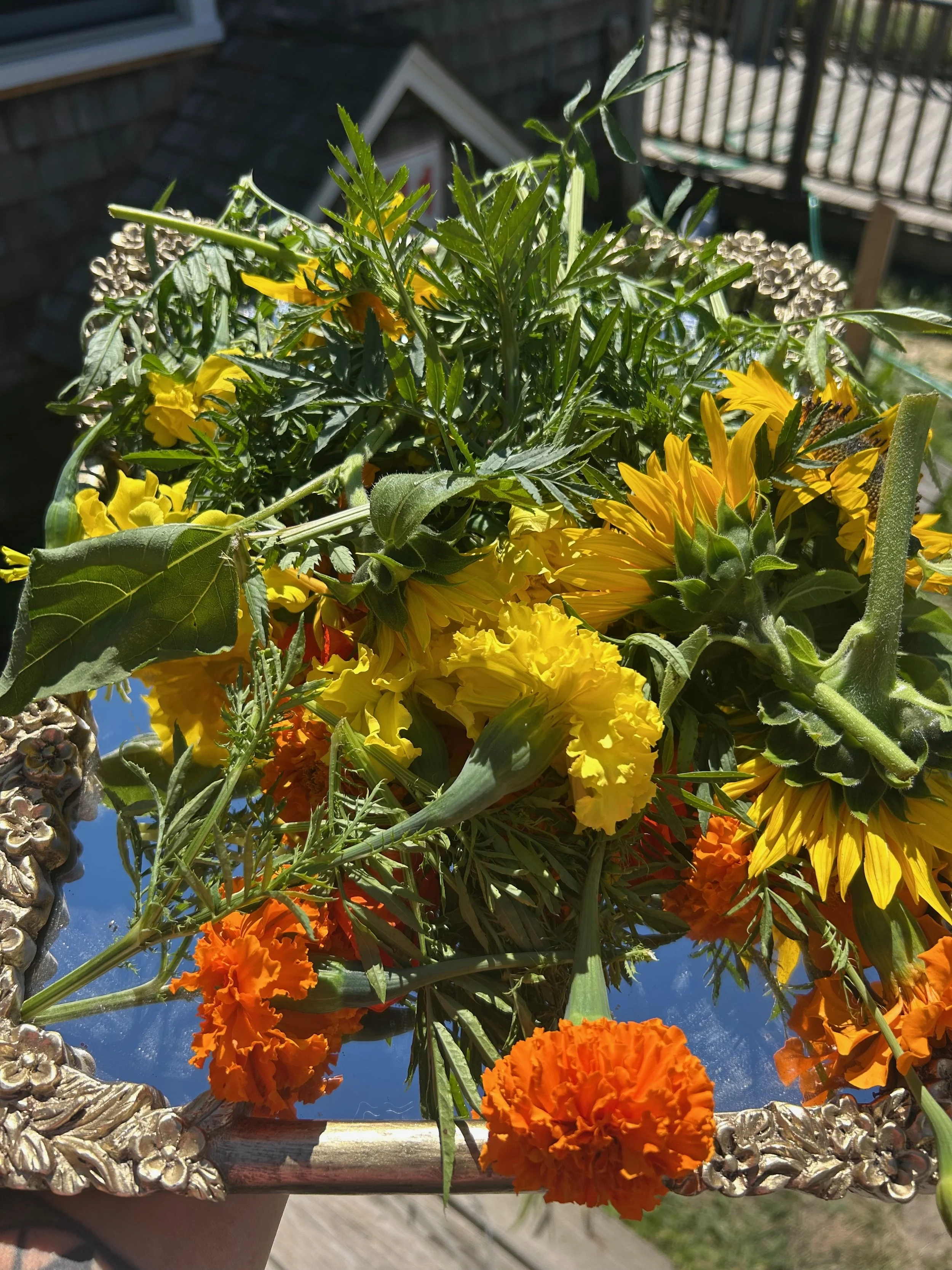 Bouquet of yellow and orange flowers, including marigolds, arranged in a decorative silver frame, outdoors with a house and fence in the background.
