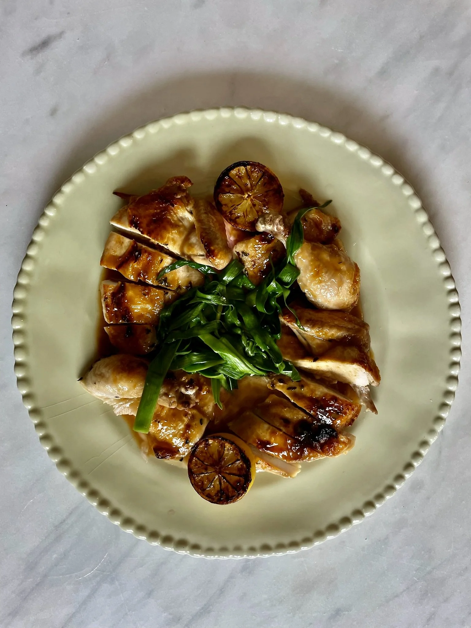 Sliced roasted chicken topped with green onions, grilled lemon slices, and sauce on a white beaded plate.