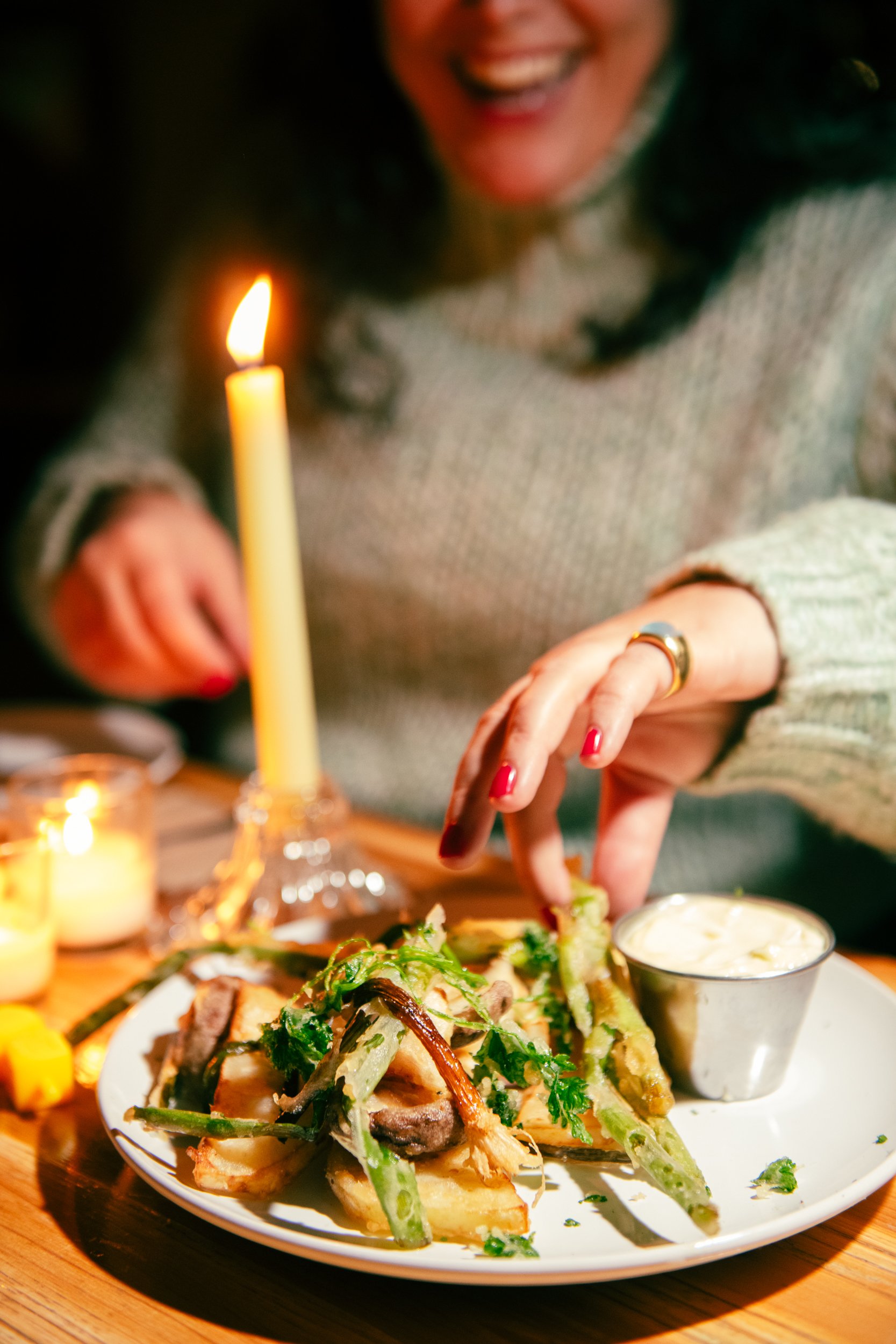 Person smiling and reaching for a plate of food with green beans and a dipping sauce at a candlelit dinner.