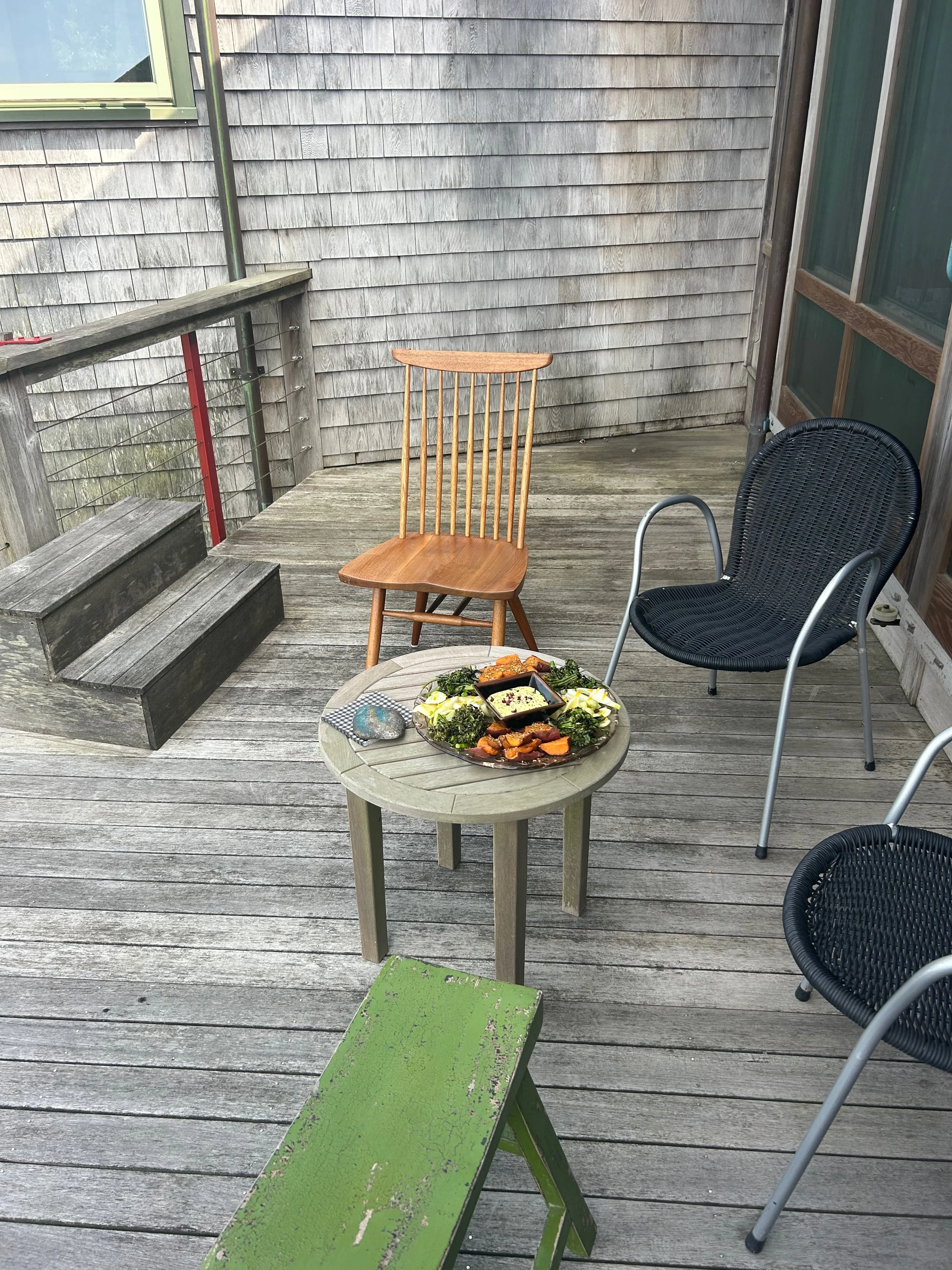 Outdoor wooden deck with three chairs and a small table holding food. Two black chairs with metal frames, one wooden chair with vertical slats, and a small green wooden bench. The table has a platter of food and condiments.