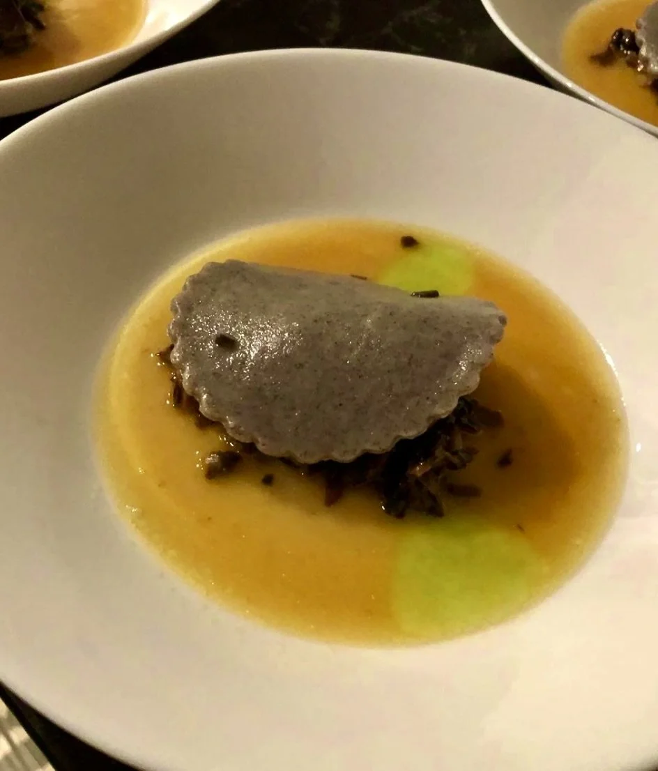 A bowl of soup with a gray dumpling on top, surrounded by yellowish broth and small dark ingredients.