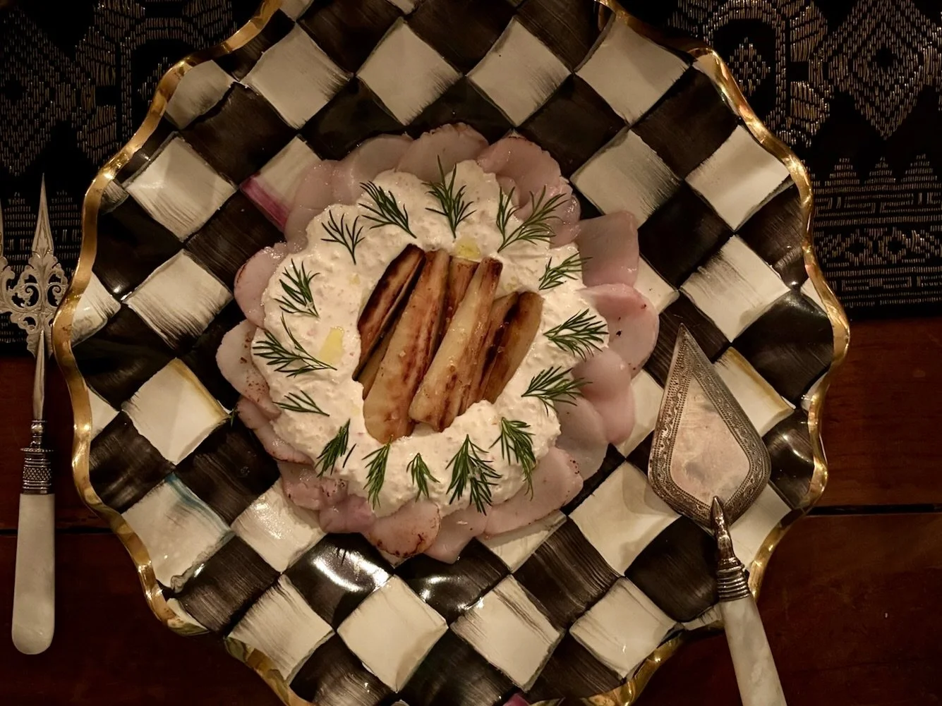 A plate of scallop crudo garnished with fresh dill, with roasted parsnip in the center, served on a McKenzie Childs. Silver serving utensils are on either side of the plate. Private Dining Martha’s Vineyard