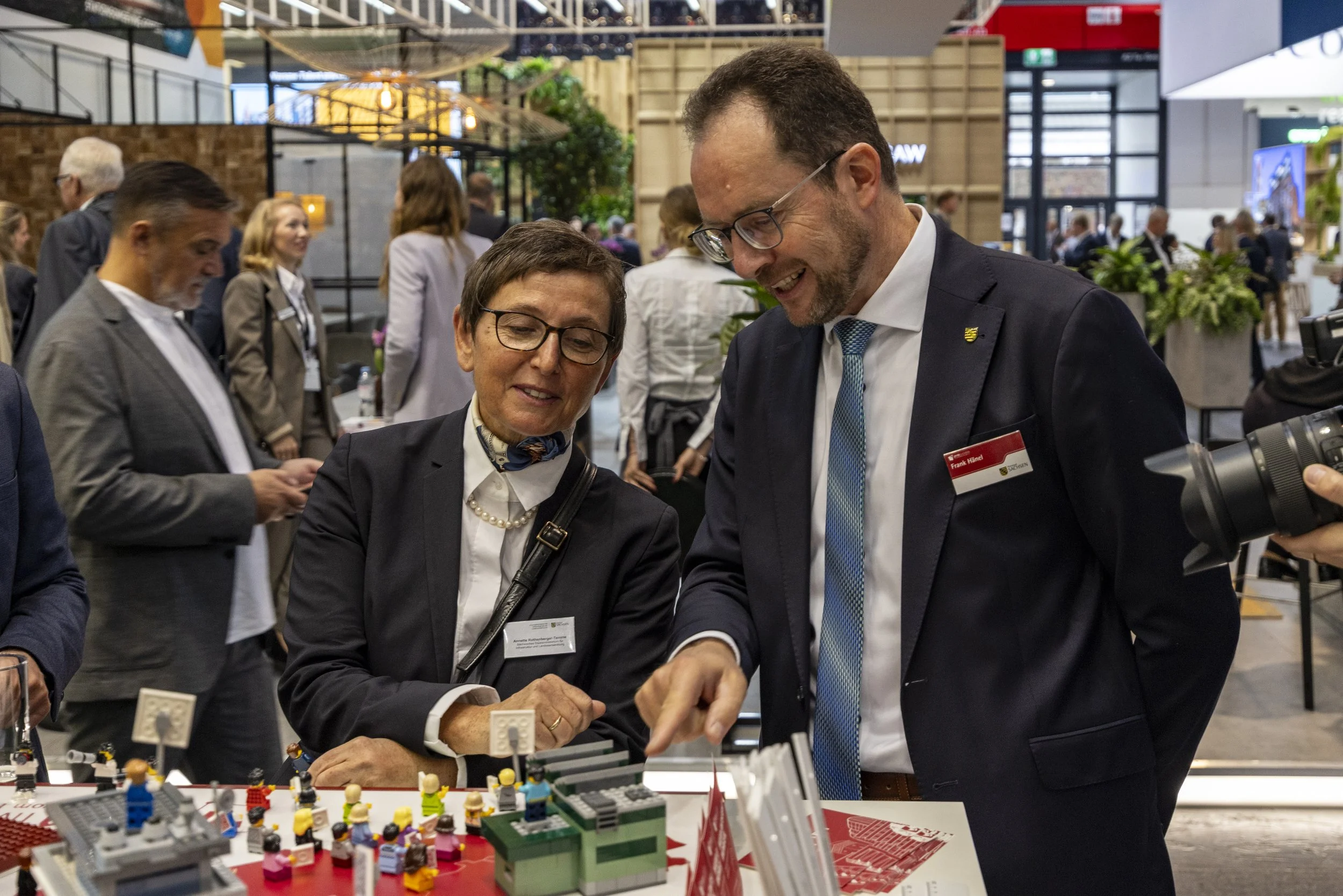 Zwei Männer im Anzug und Brille schauen auf eine Lego-Ausstellung mit Lego-Figuren, während im Hintergrund weitere Menschen im Gespräch sind.