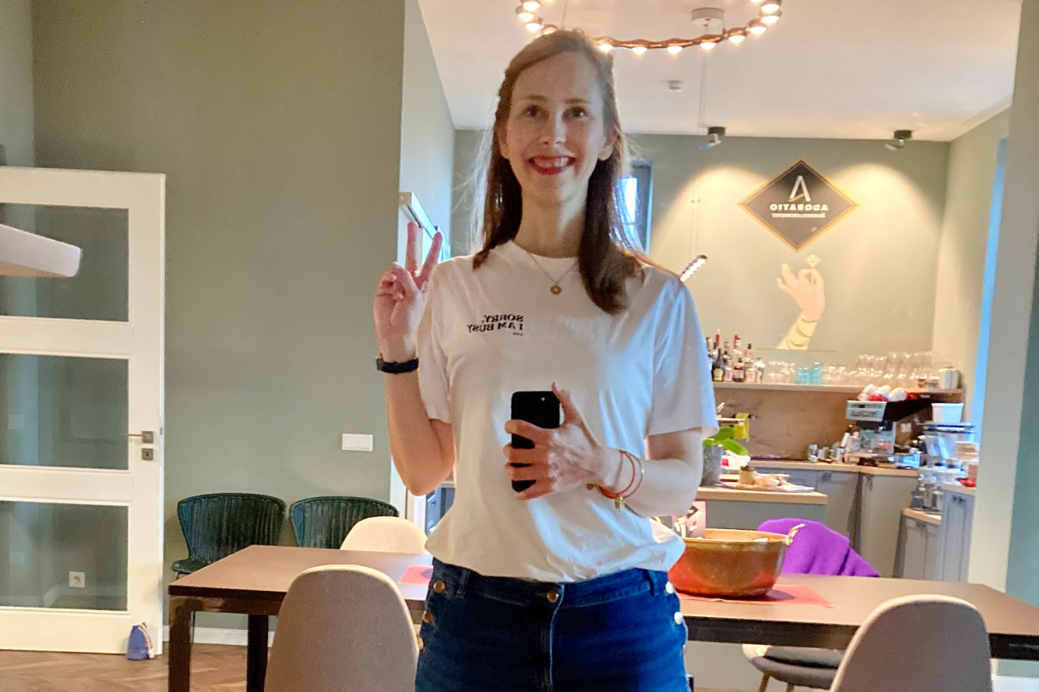 Junge Frau macht ein Selfie im Wohnzimmer, trägt ein weißes T-Shirt und Jeans, zeigt den peace-Zeichen und lächelt.