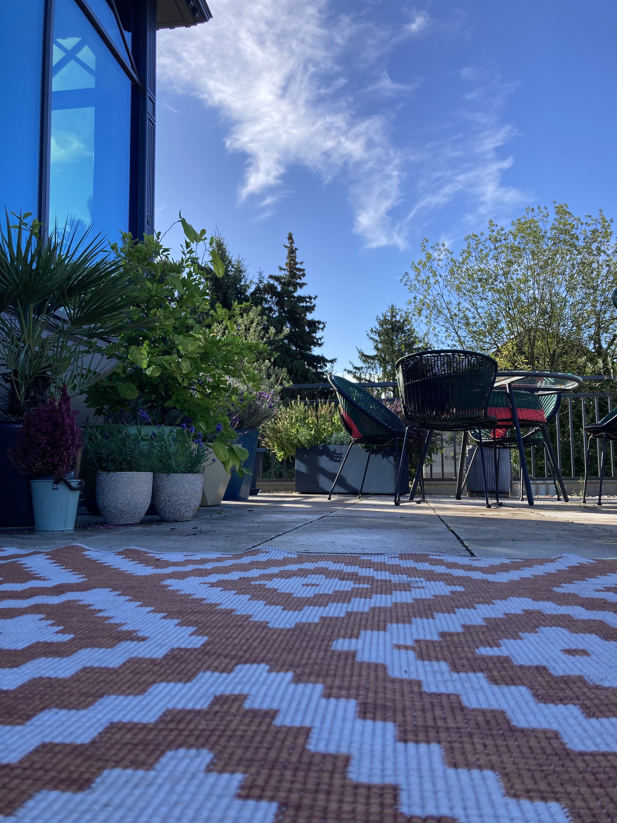 Blick auf einen Balkon mit mehreren Topfpflanzen und Gartenmöbeln, darunter Stühle und ein Tisch, bei schönem Wetter mit blauen Himmel und Wolken.