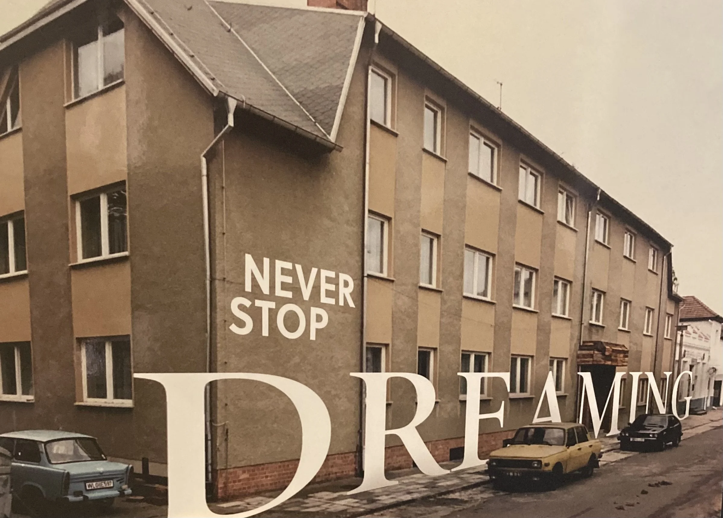 Ein Mehrfamilienhaus mit mehreren Fenstern und einer großen Werbetafel an der Seite, auf der steht 'NEVER STOP DREAMING'. Es sind mehrere geparkte Autos vor dem Gebäude zu sehen.