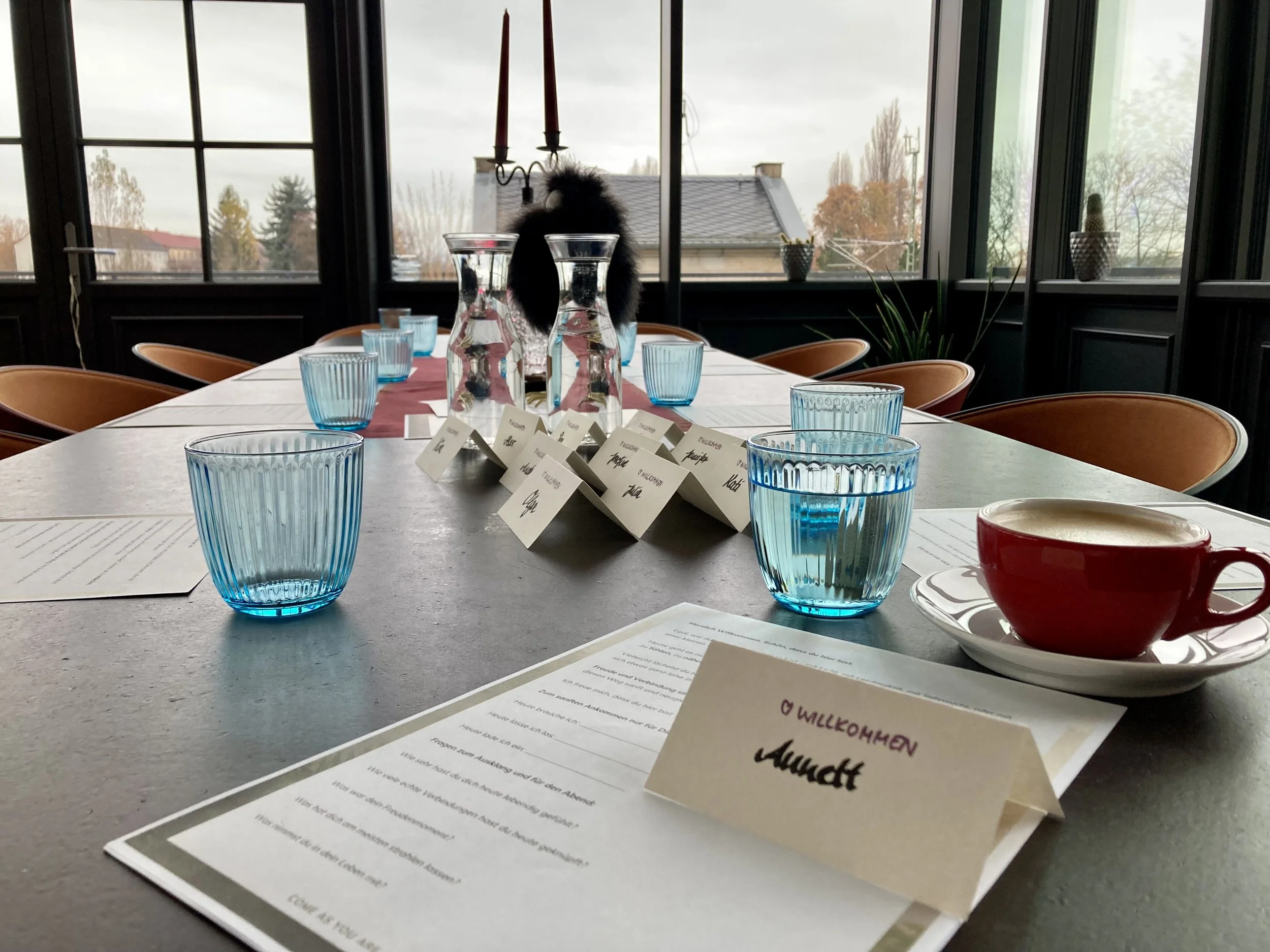 Ein Esstisch mit blauen Gläsern, Menükarten und einer roten Kaffeetasse vor großen Fenstern mit Blick auf ein Dach und Bäume im Herbst. Im Hintergrund stehen zwei Kerzen auf einem Kerzenhalter.